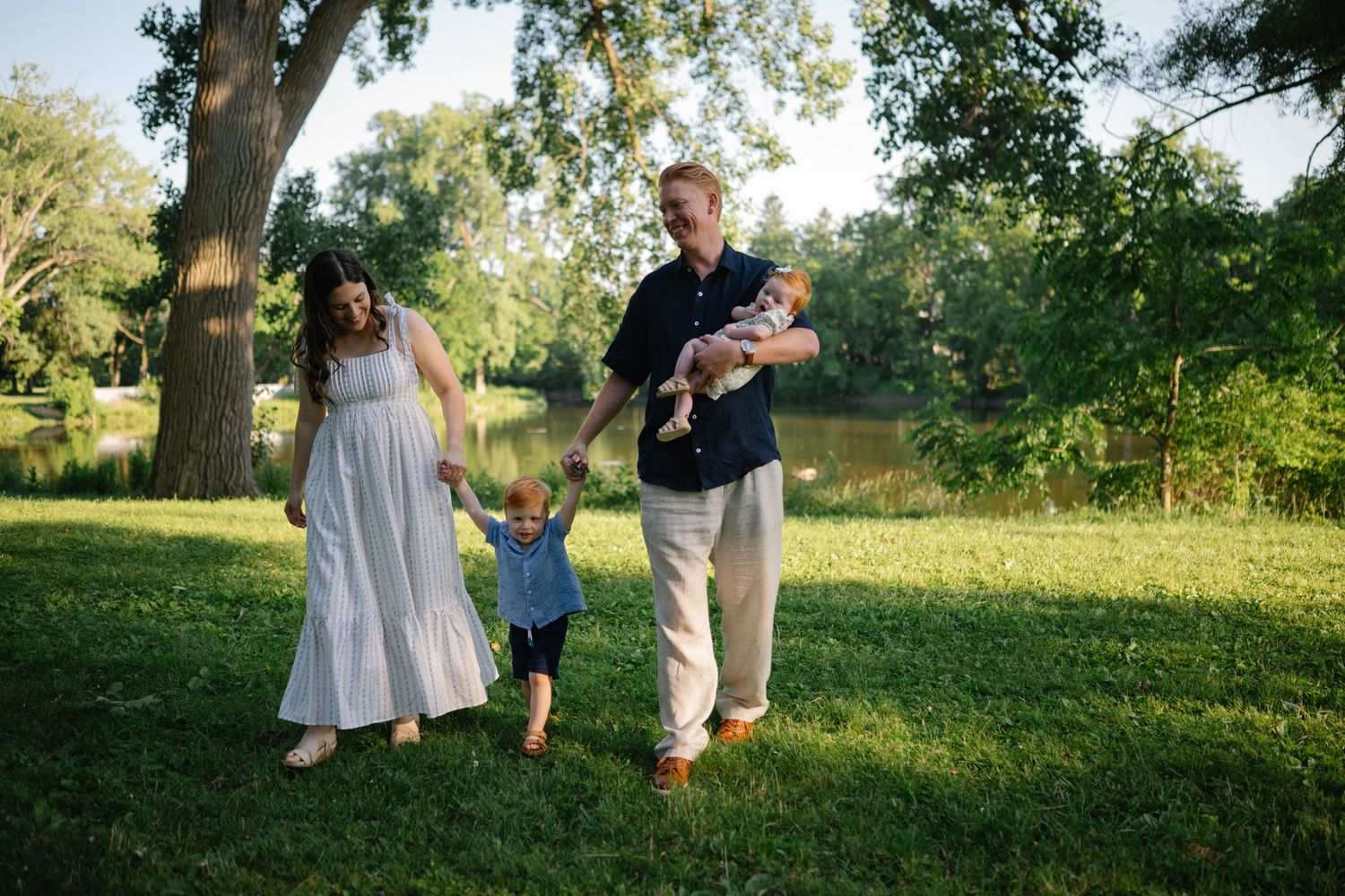 Family of four walking outdoors near a pond, parents holding children's hands on a sunny day in a lush green park.