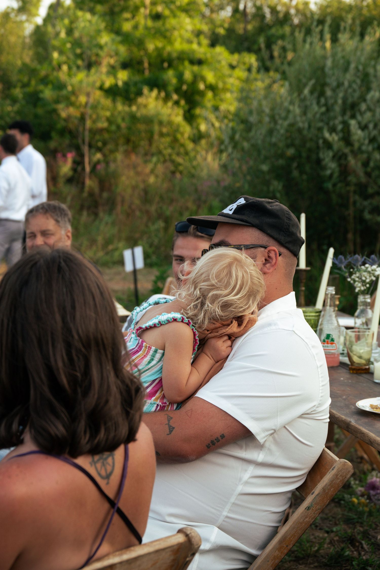 Man in white shirt and black cap holds a young child at an outdoor garden gathering with family.