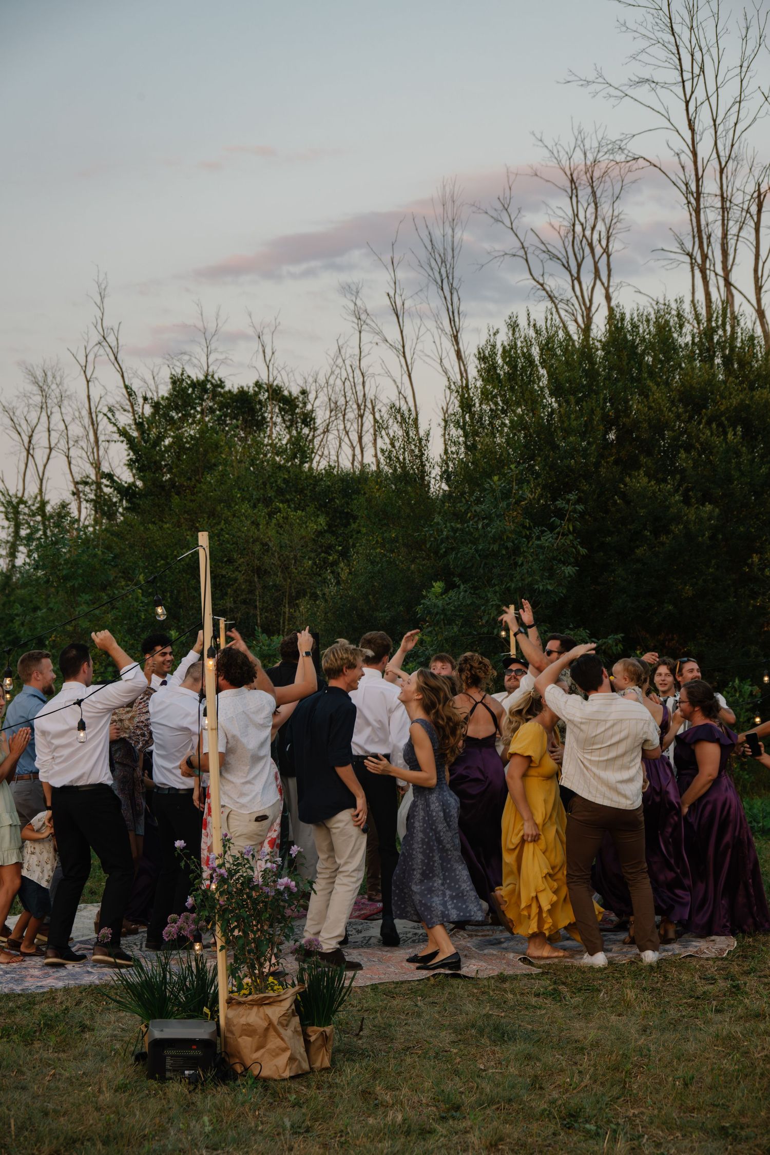 Guests dancing outdoors at a wedding reception during golden hour, surrounded by lush green trees and colorful attire.