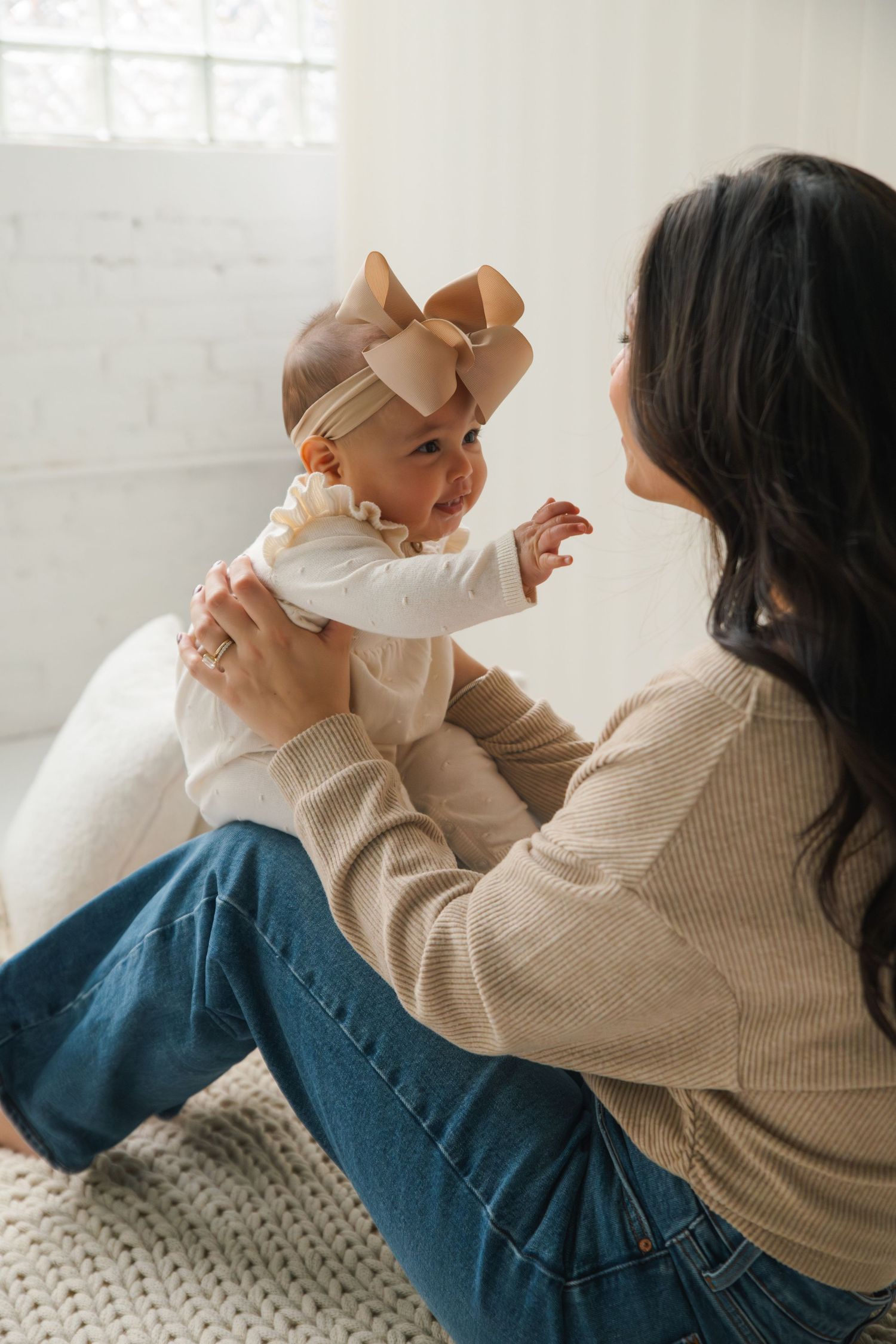 Parent in beige sweater and blue jeans holds and plays with baby wearing light colored outfit on white bed.