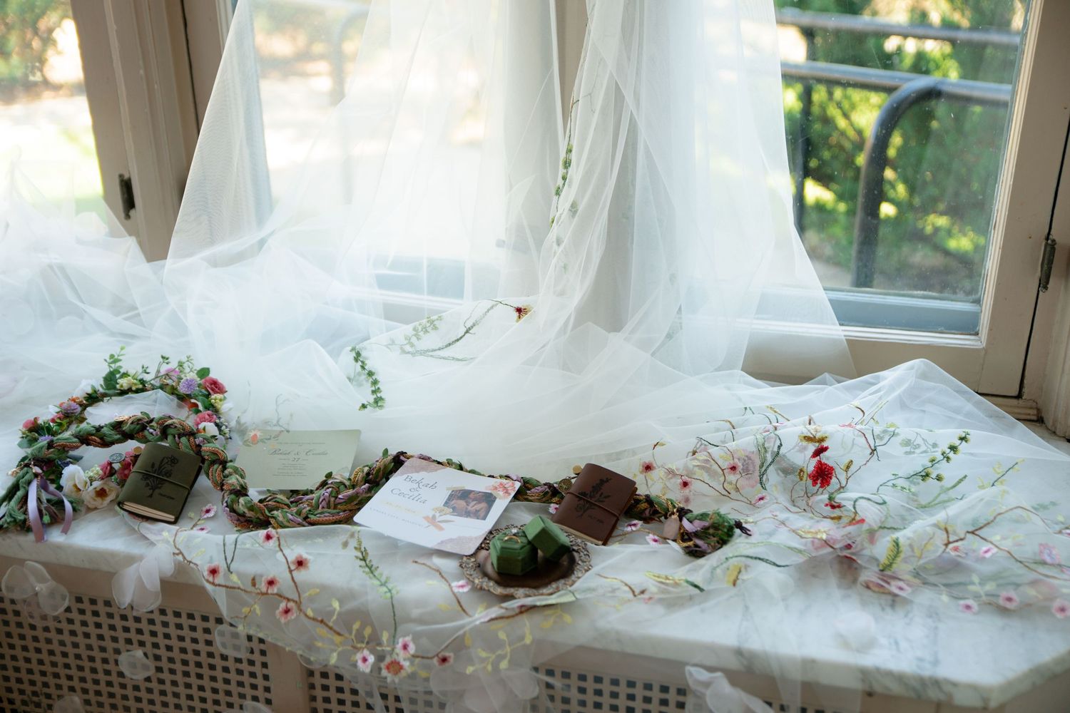 Elegant wedding flat lay with floral crown, veil, and stationery arranged on a white table by a sunlit window.