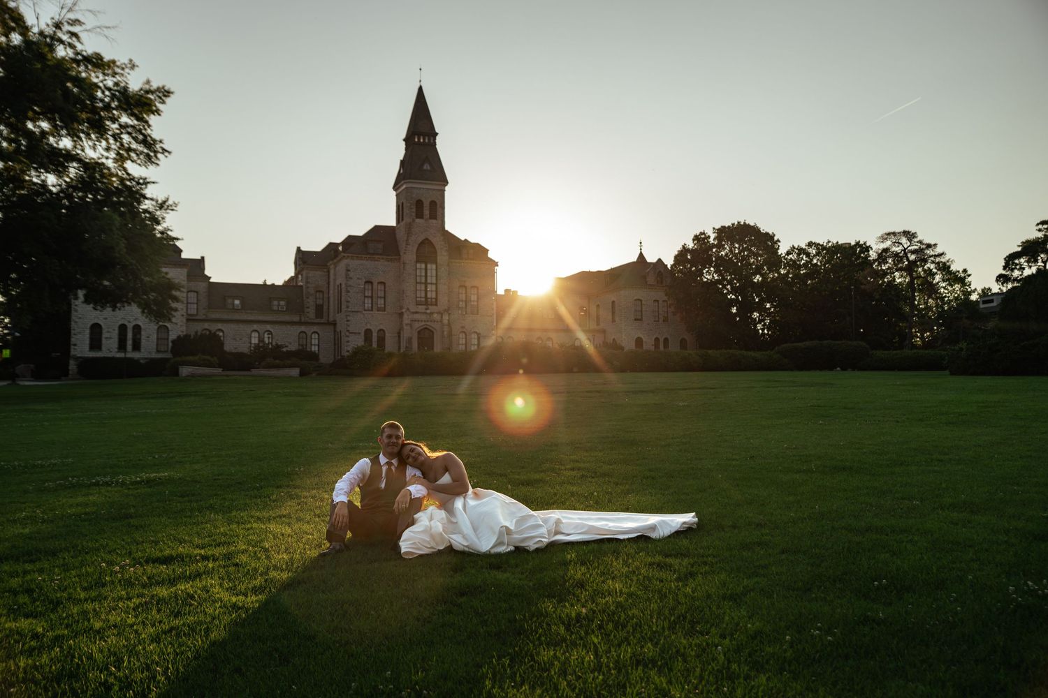 Bride and groom sitting on lush green lawn at sunset with historic stone building in background.