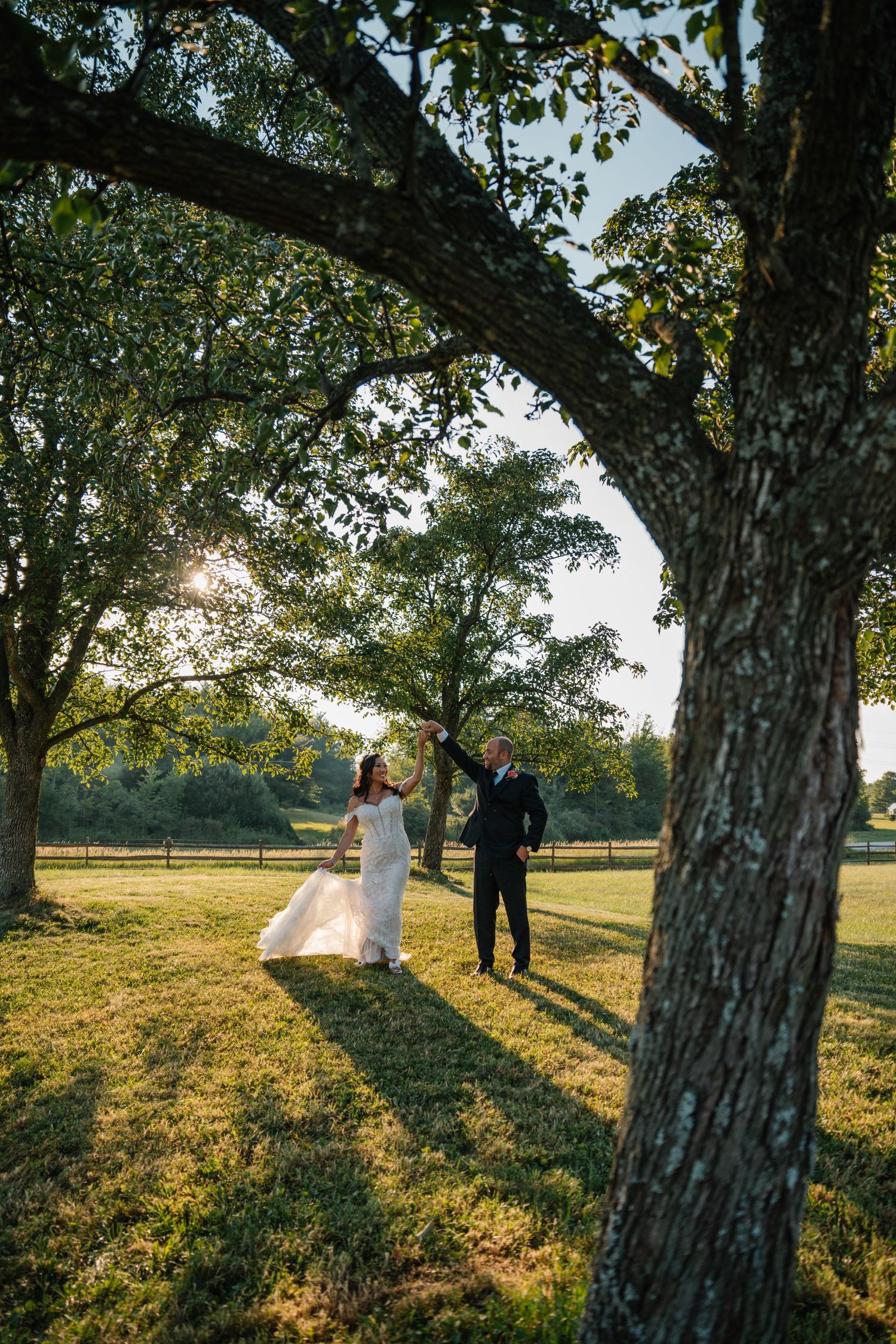 Bride and groom dancing under large trees in a sunlit green field during golden hour outdoor wedding.