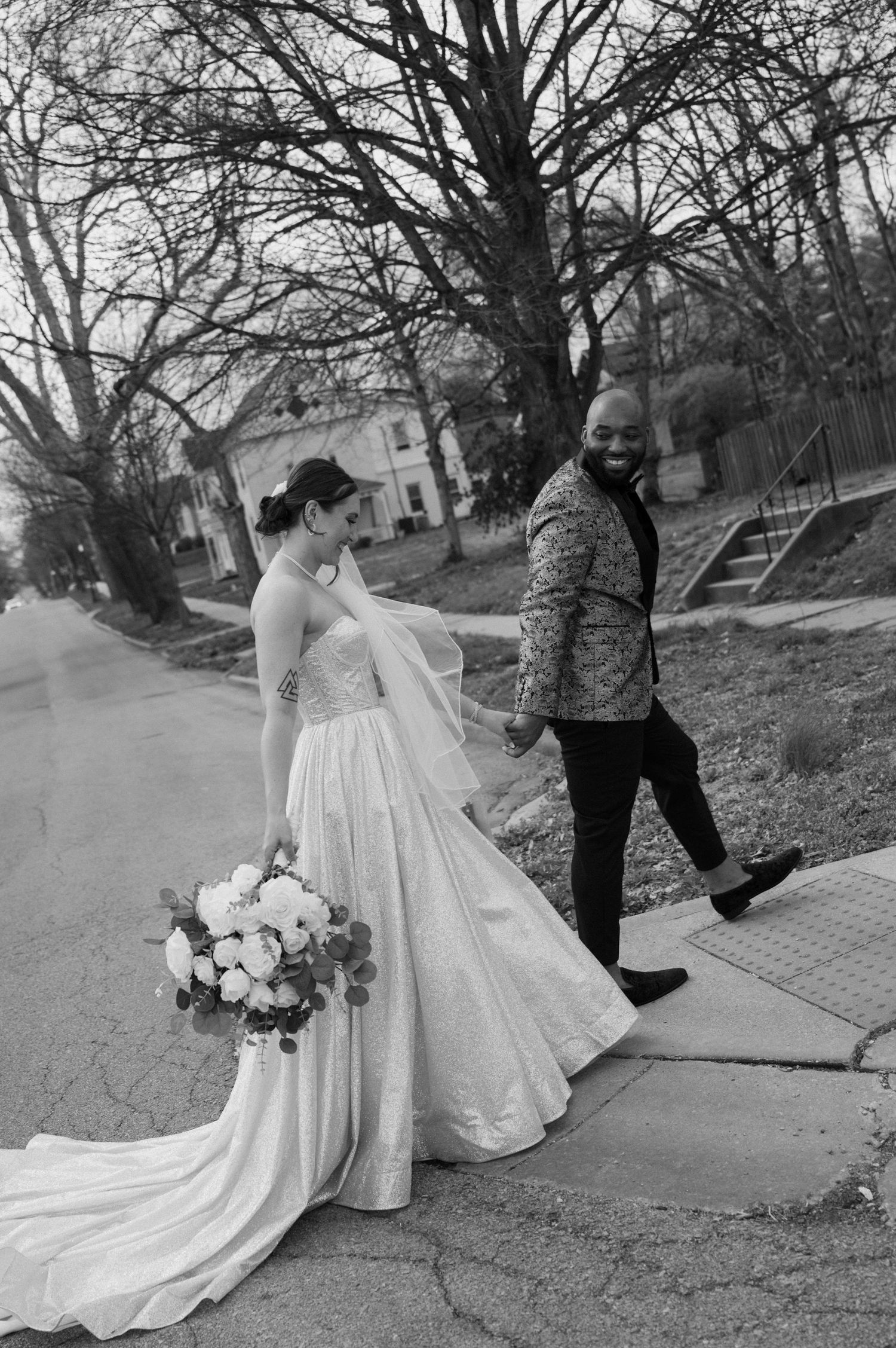 A bride holding a floral bouquet walks with her partner on a suburban street in black and white.