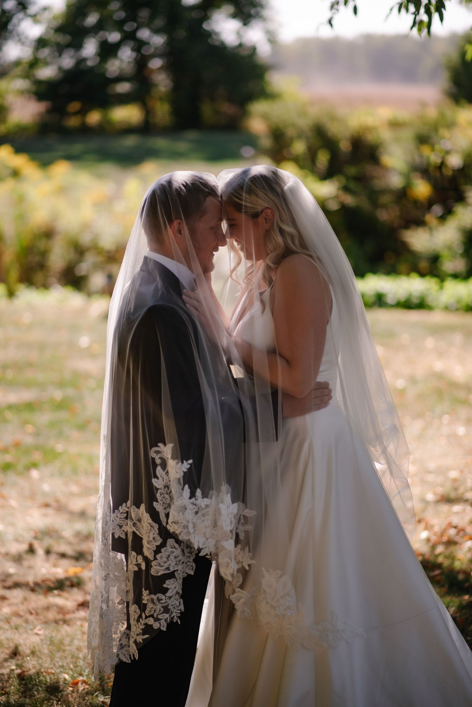 Bride and groom share an intimate moment under a lace-trimmed veil in a sunlit garden setting.