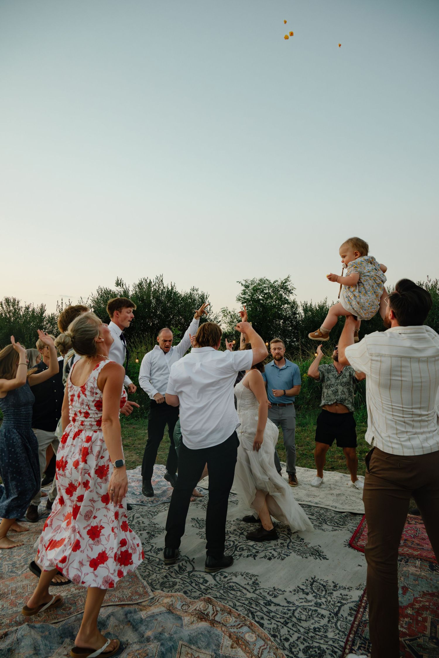 Wedding guests celebrate outdoors at dusk, dancing on a rug while a child is lifted joyfully into the air.