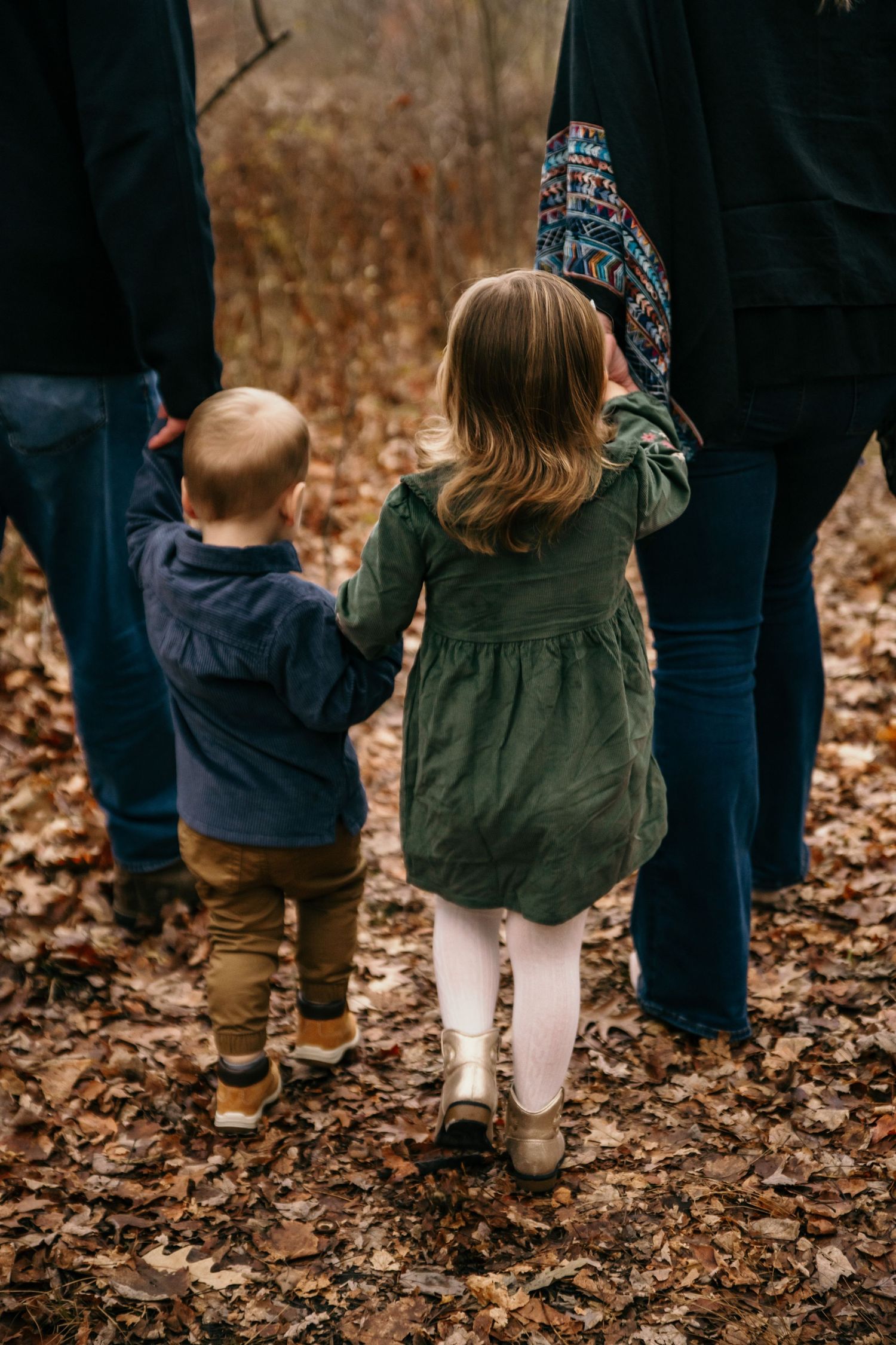 Two children hold hands with adults while walking through autumn leaves.