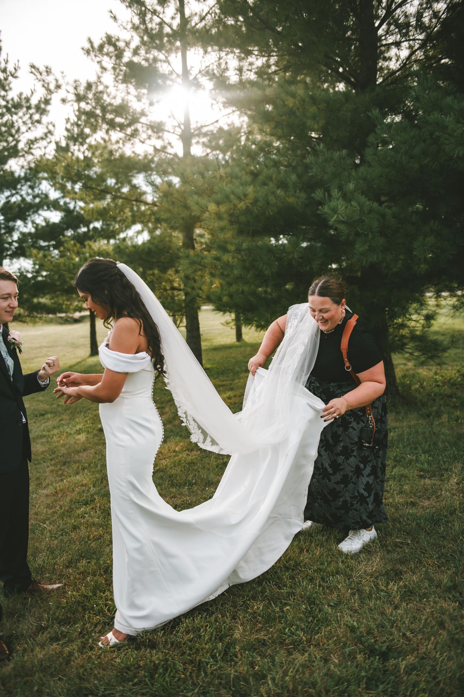 Wedding party playfully posing with bride's dress in a grassy field at sunset.