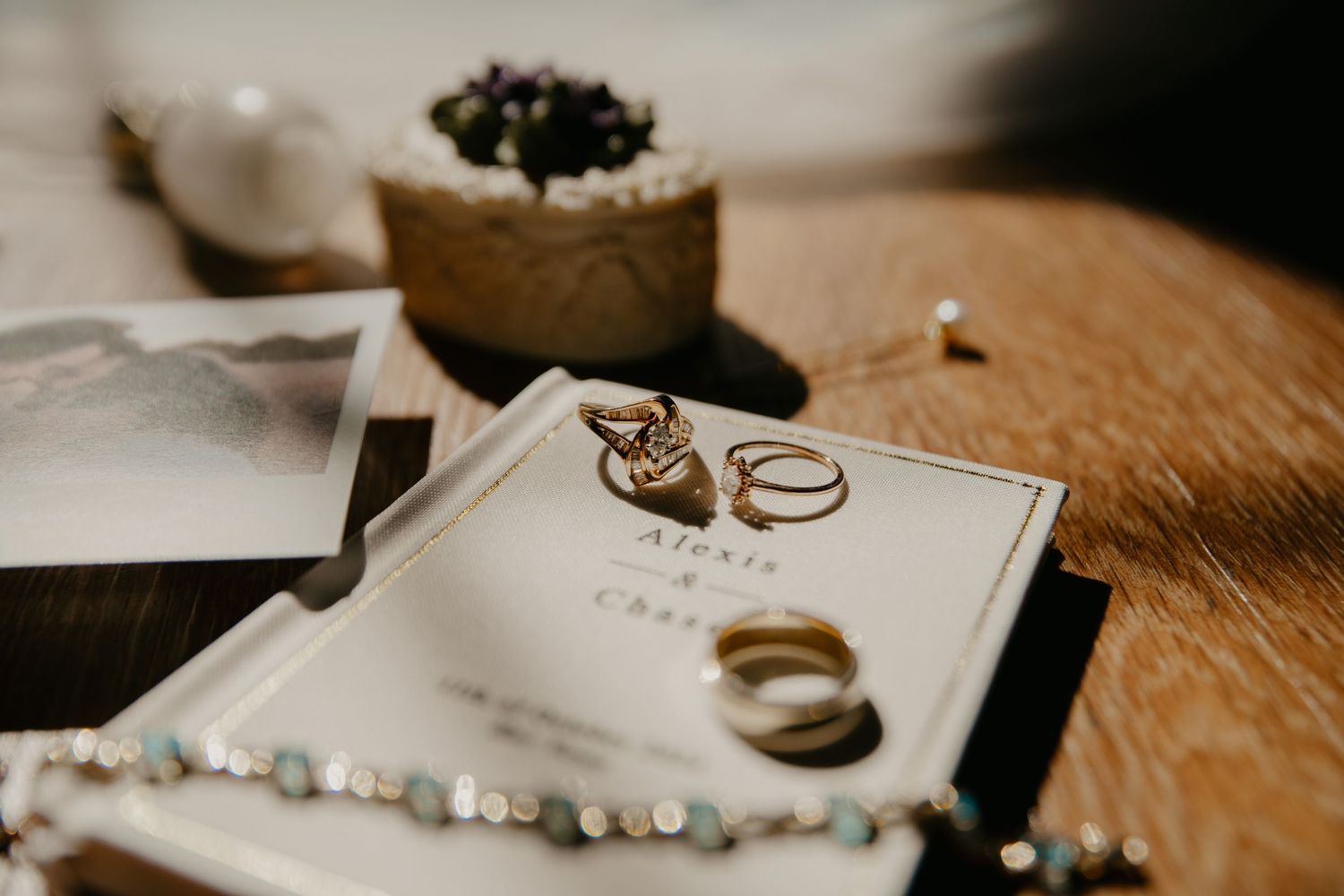 Gold wedding rings and bands rest on an elegant wedding invitation beside pearl jewelry on a wooden table.