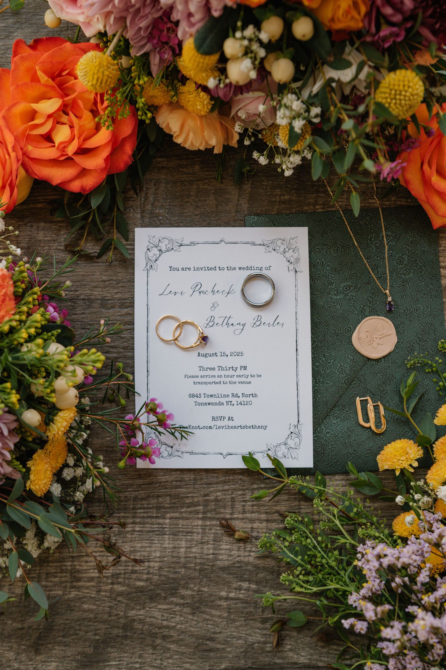 Wedding invitation for Lora and Brittany surrounded by colorful wildflowers, gold rings, and elegant jewelry on rustic wood.