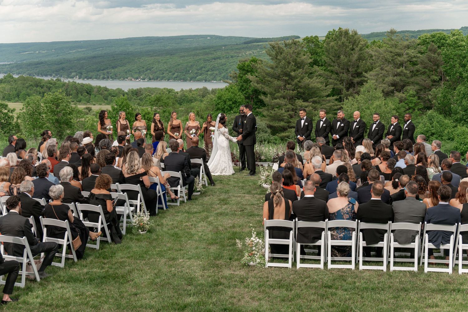 Outdoor wedding ceremony on a hillside with guests seated in white chairs, overlooking a scenic lake and green landscape.