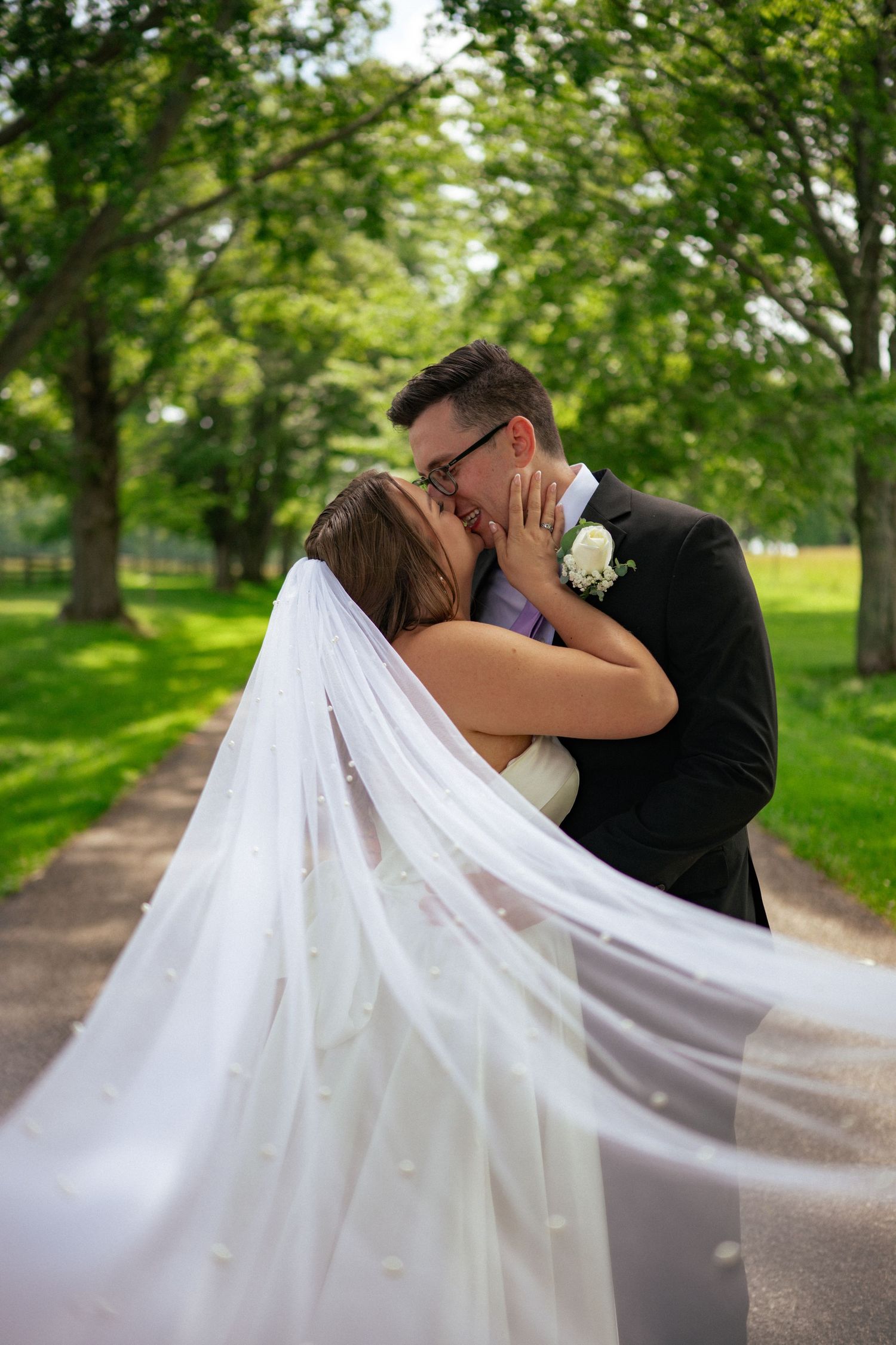 Bride and groom share a romantic kiss outdoors, with her long pearl-adorned veil flowing in the breeze amid lush green trees.