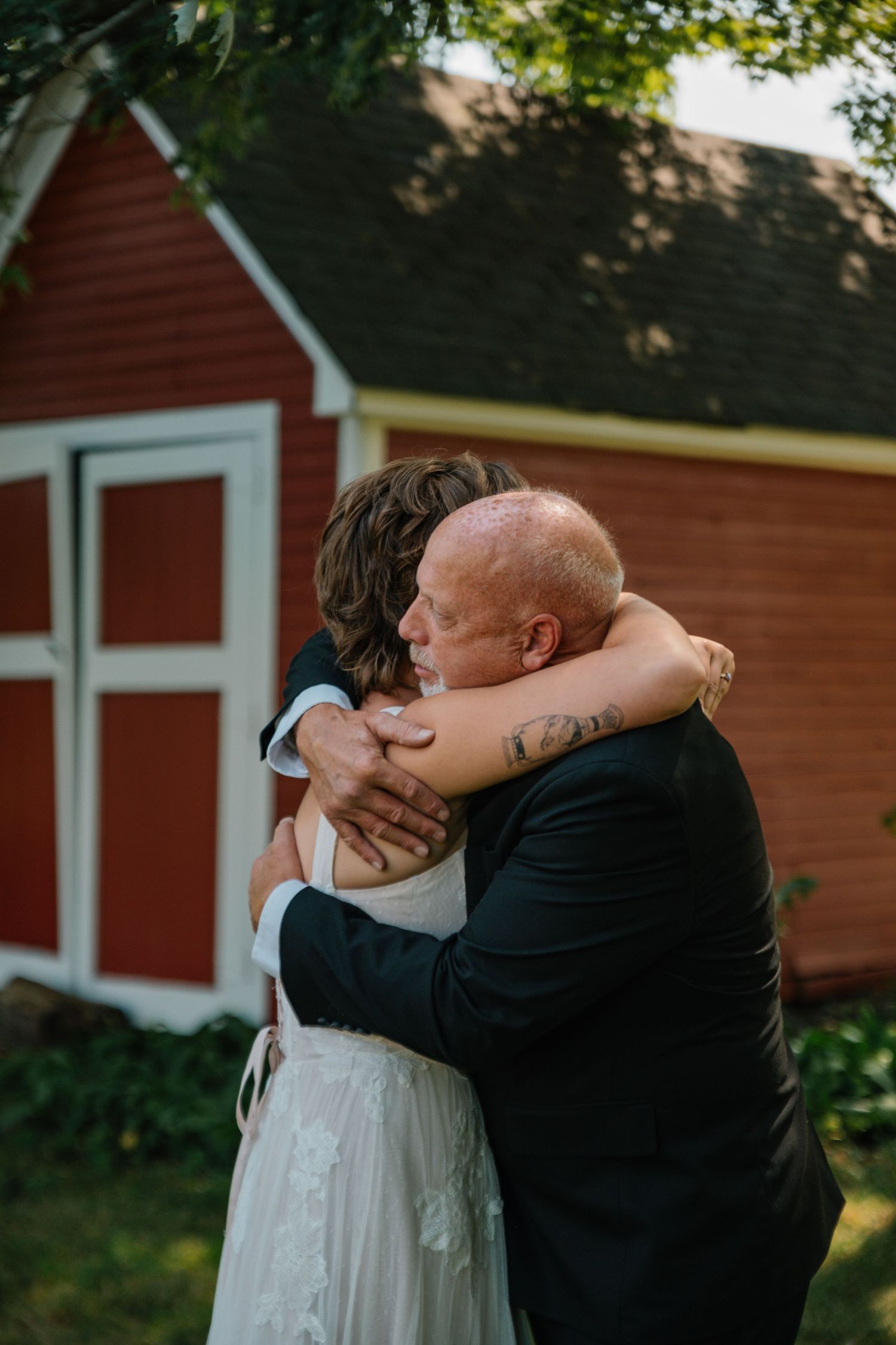 A bride and older man share an emotional hug outdoors near a red barn during a wedding celebration.