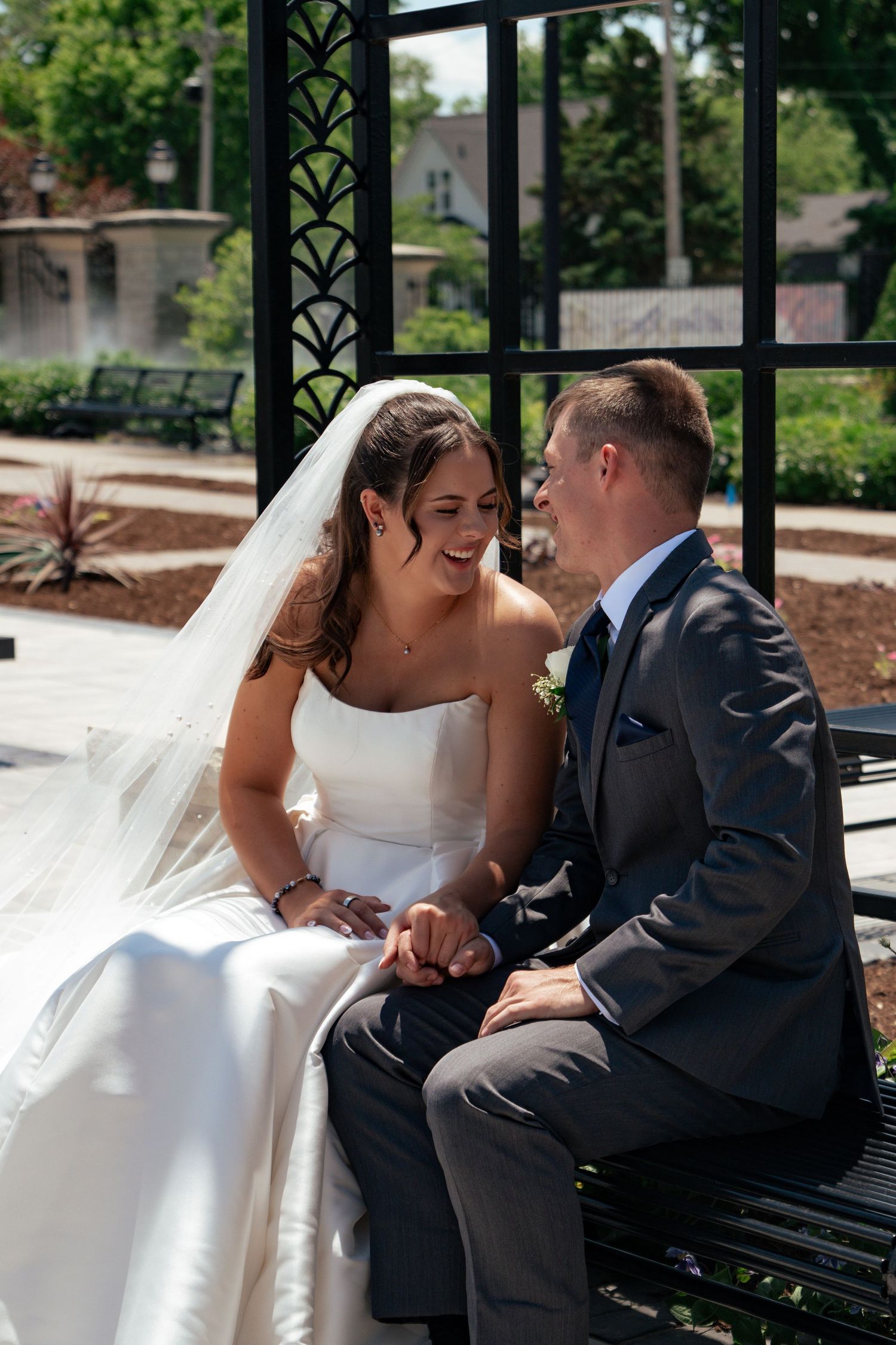 A smiling bride in white gown and veil holds hands with groom in gray suit on a bench in a sunny garden.