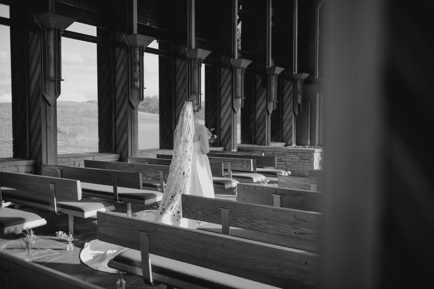 A wedding veil hangs between wooden church pews in a dramatic black and white composition.