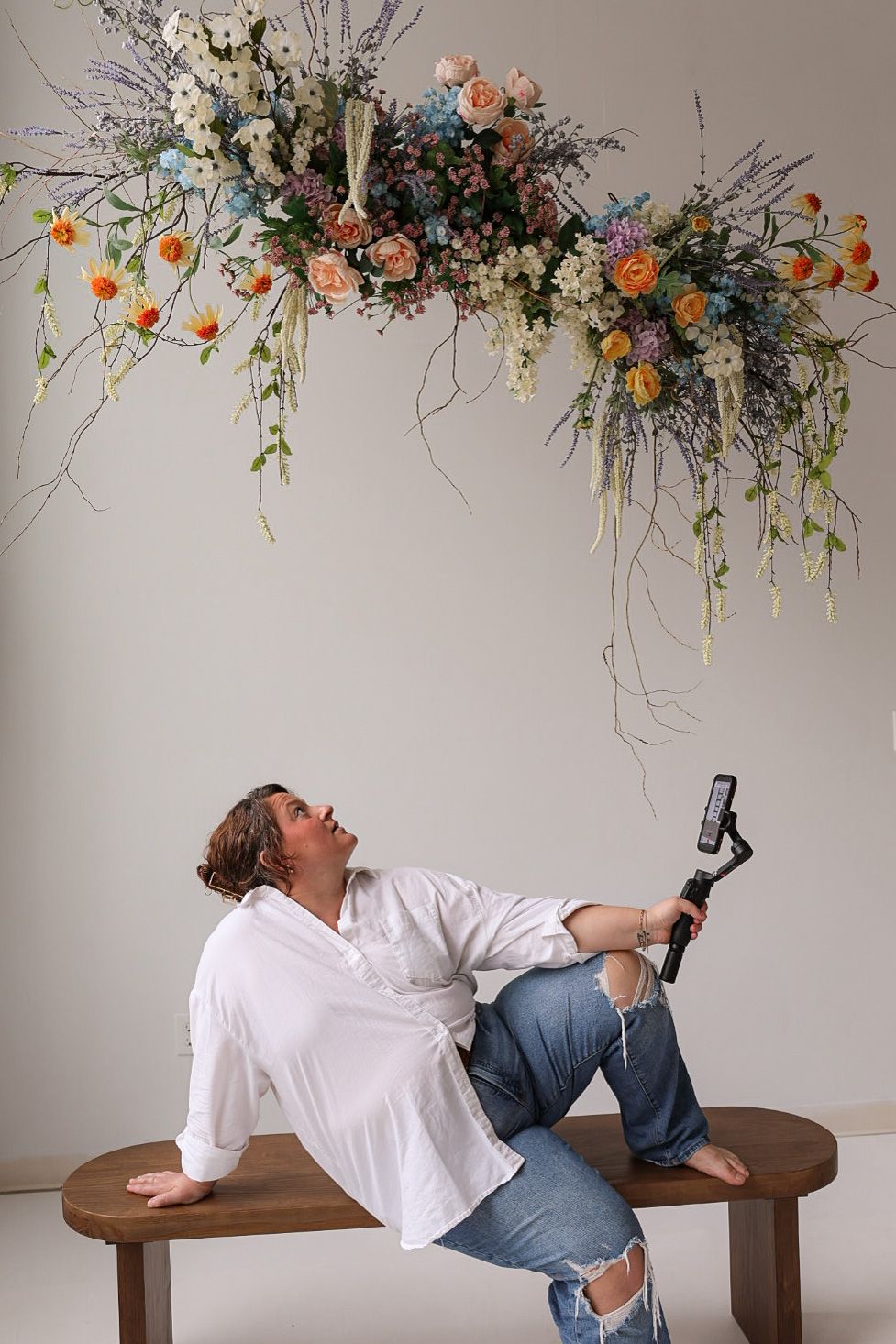 Person in white shirt and ripped jeans reclines on bench, filming overhead floral arrangement with phone gimbal.