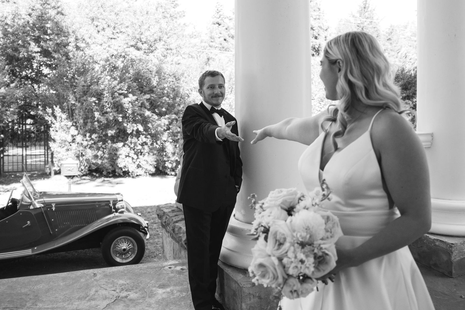 Black and white wedding photo of bride holding bouquet and groom in tuxedo near vintage car and columns.