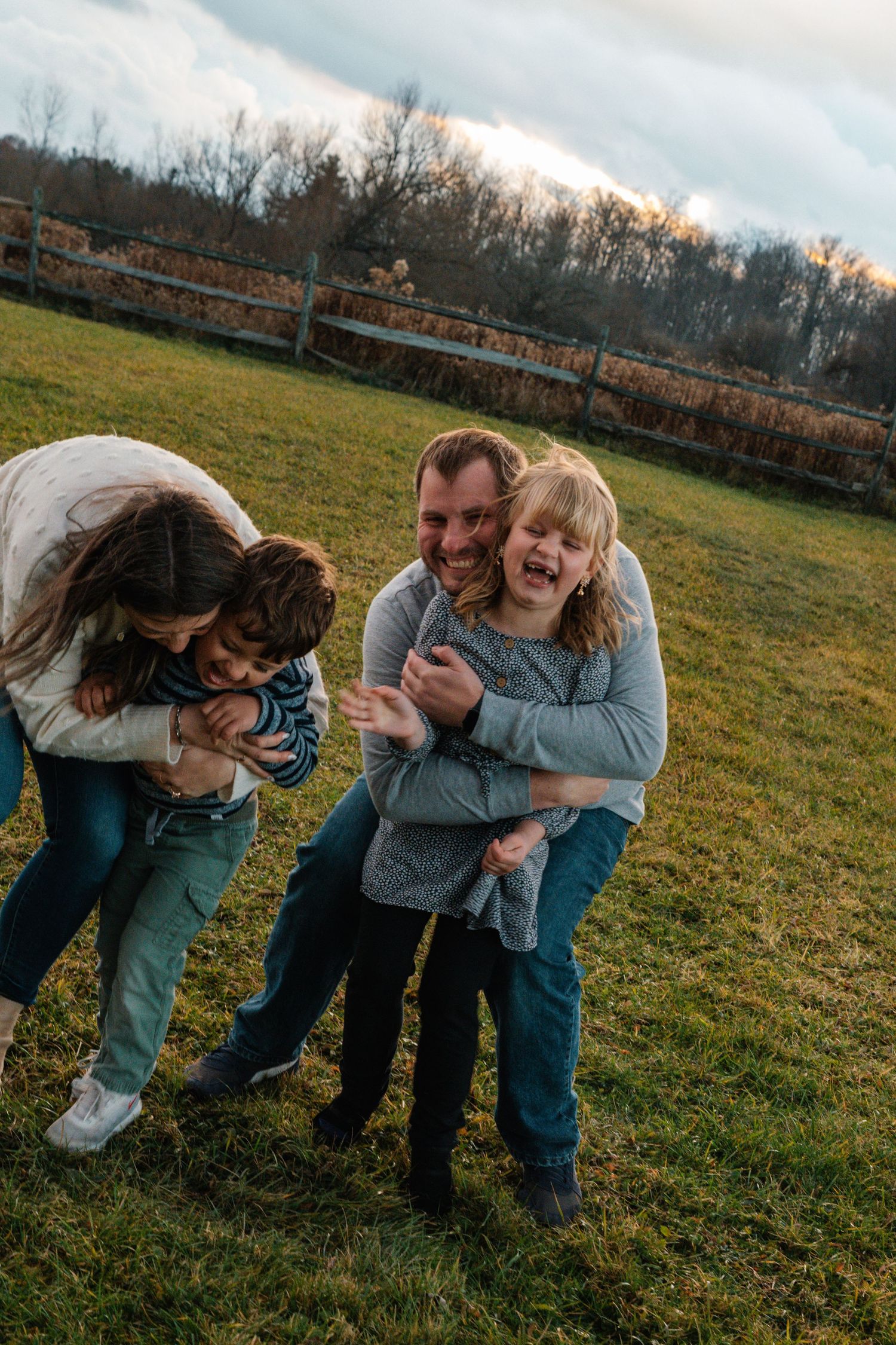 A joyful family moment captured outdoors in a grassy field with a wooden fence in the background at sunset.