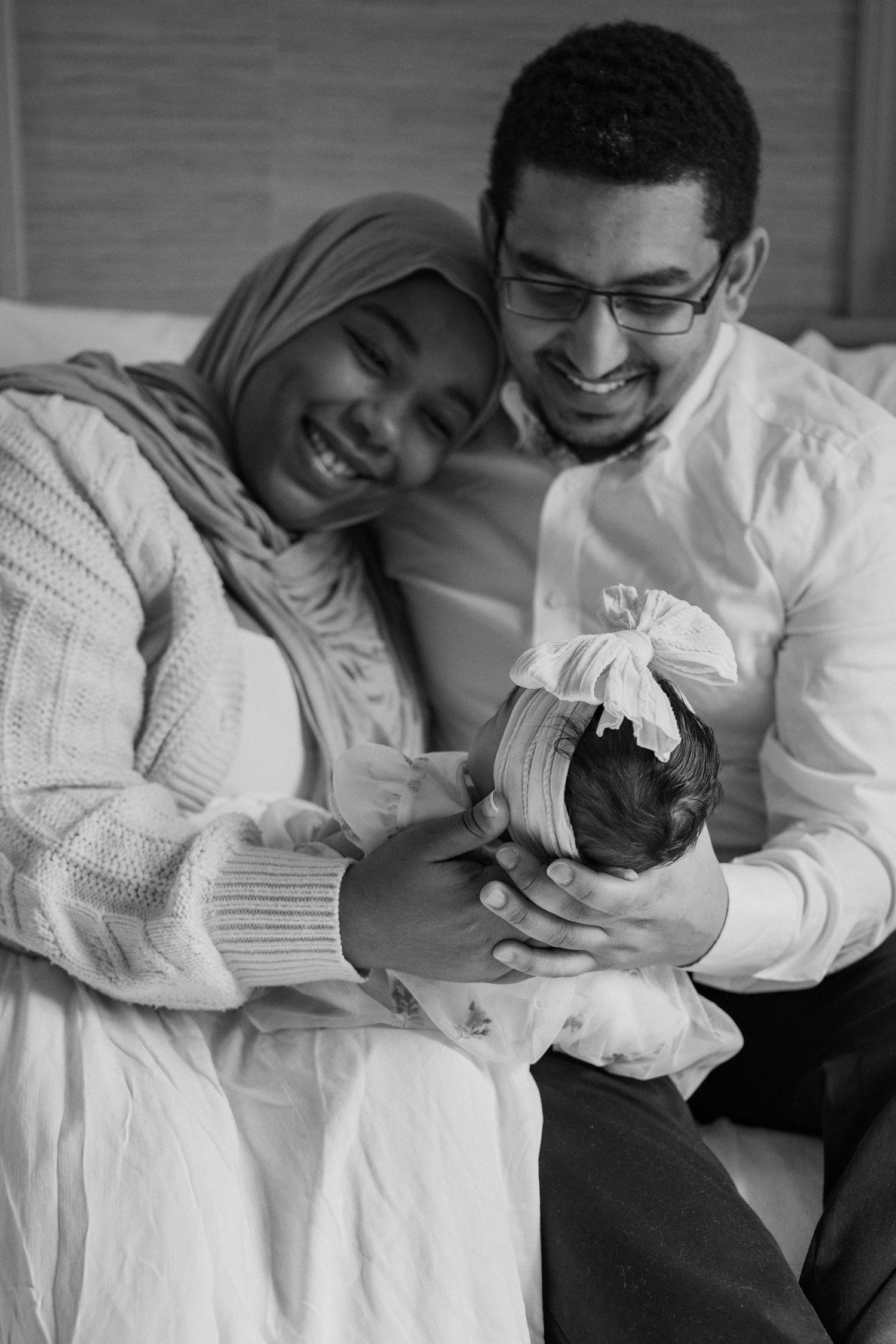 Black and white photo of smiling parents holding their newborn baby with a bow headband.