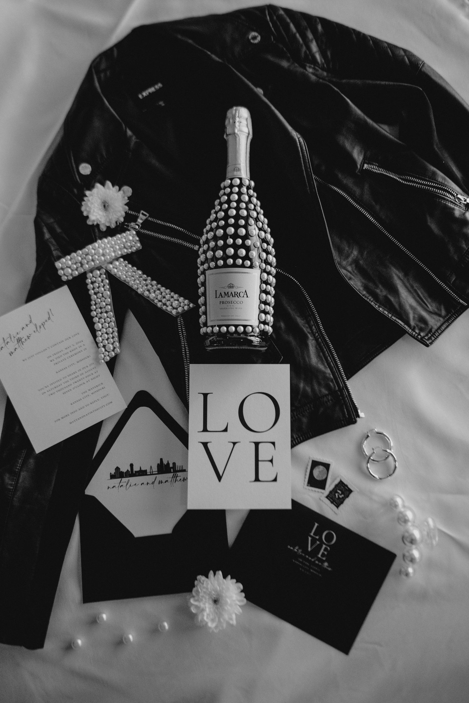 Black and white wedding details with champagne bottle, LOVE sign, rings, flowers and invitation cards on fabric background.