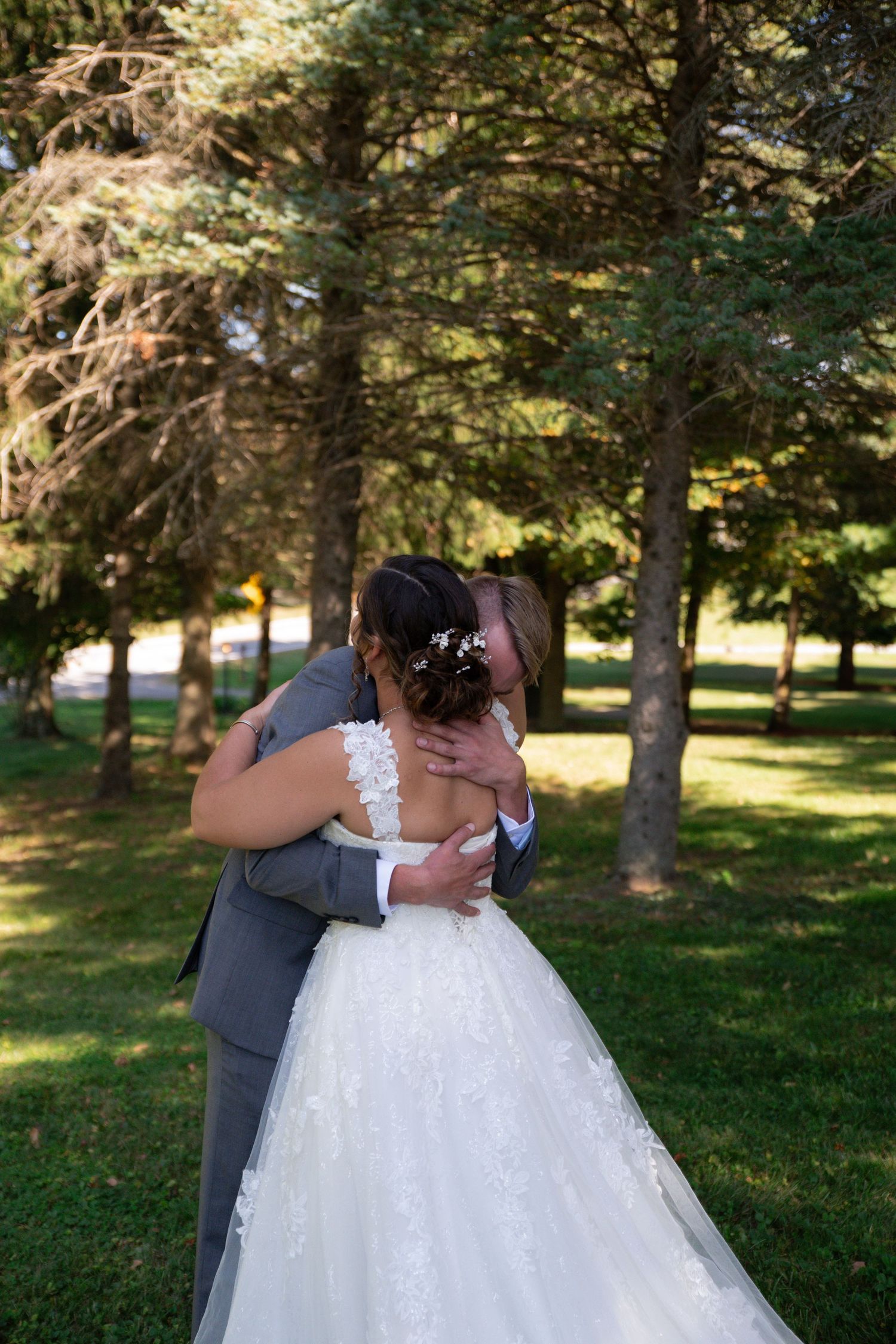 Bride and groom embracing in a lush green park, wearing a white lace gown and gray suit on their wedding day.
