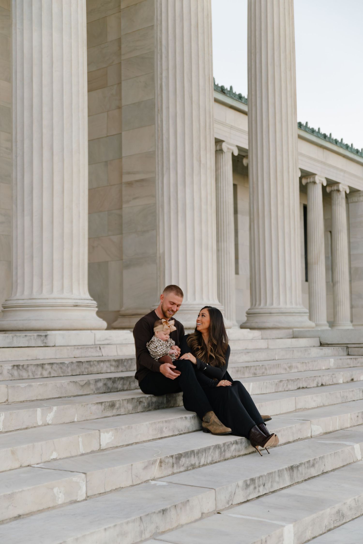 A couple sits with their dog on marble steps between classic white stone columns of a grand building.
