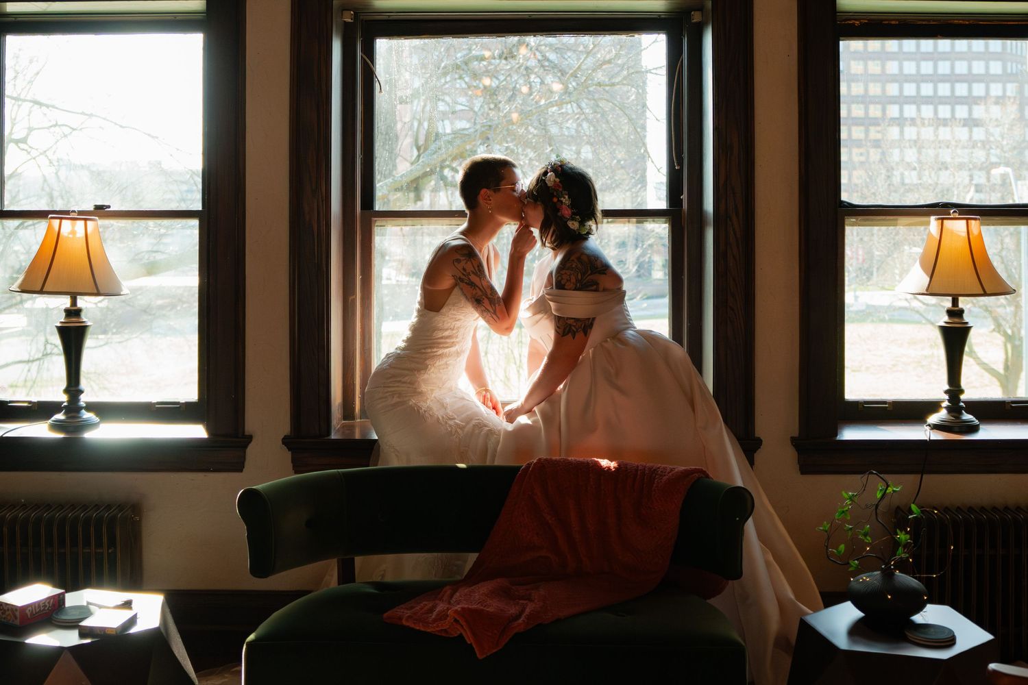 Two brides in white wedding gowns share a romantic kiss by sunlit windows with warm lamp light in an elegant room.