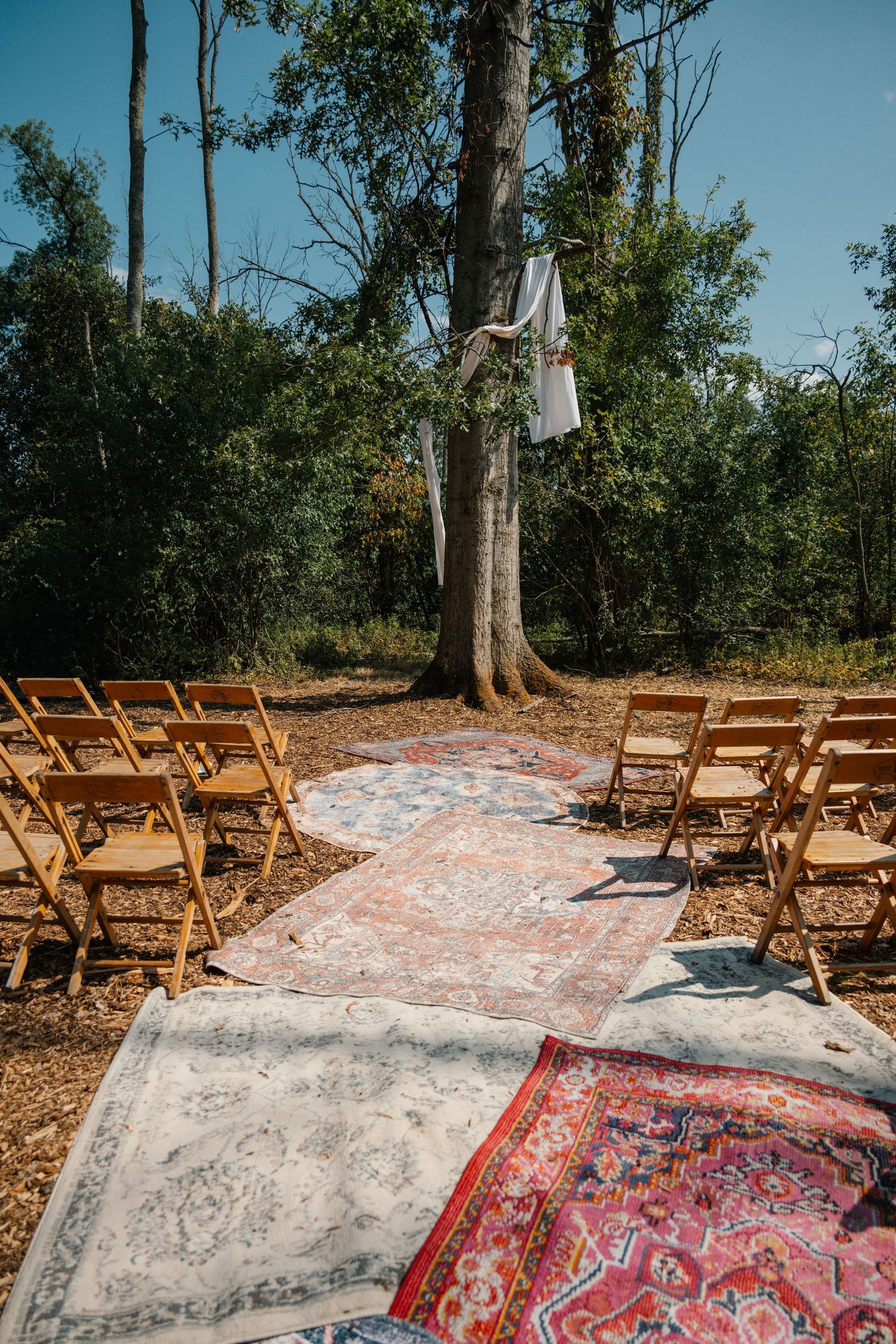 Outdoor wedding ceremony aisle with colorful vintage rugs, wooden folding chairs, and fabric hanging from a tree in a forest setting.