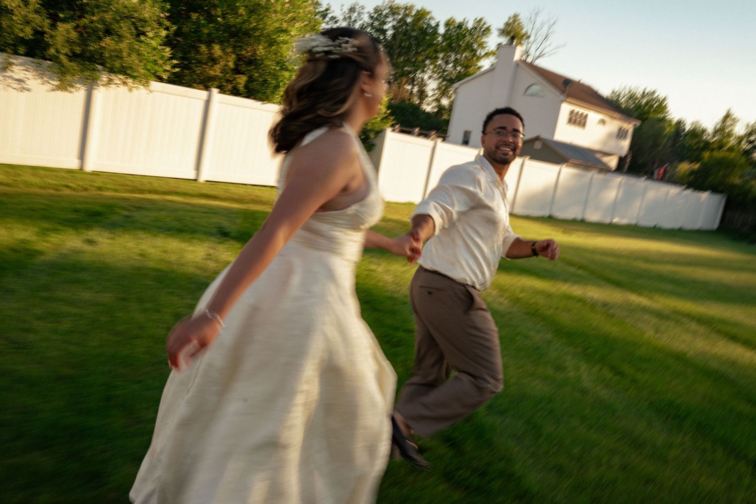 A bride in a white gown and groom in casual attire joyfully run together across a lush green backyard.