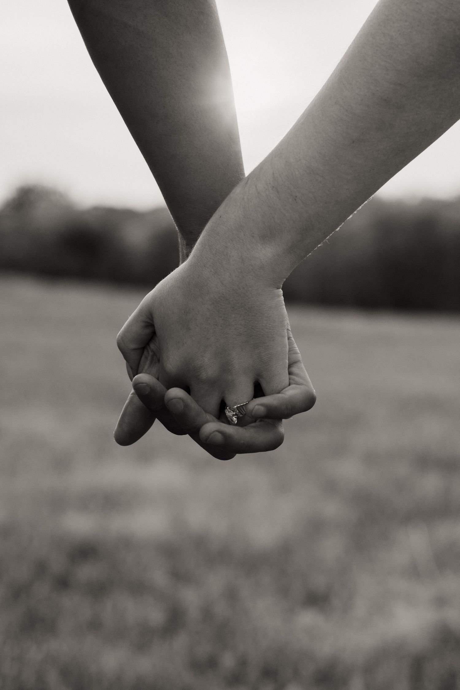 A black and white close-up photograph of two people holding hands in a field with soft bokeh background.