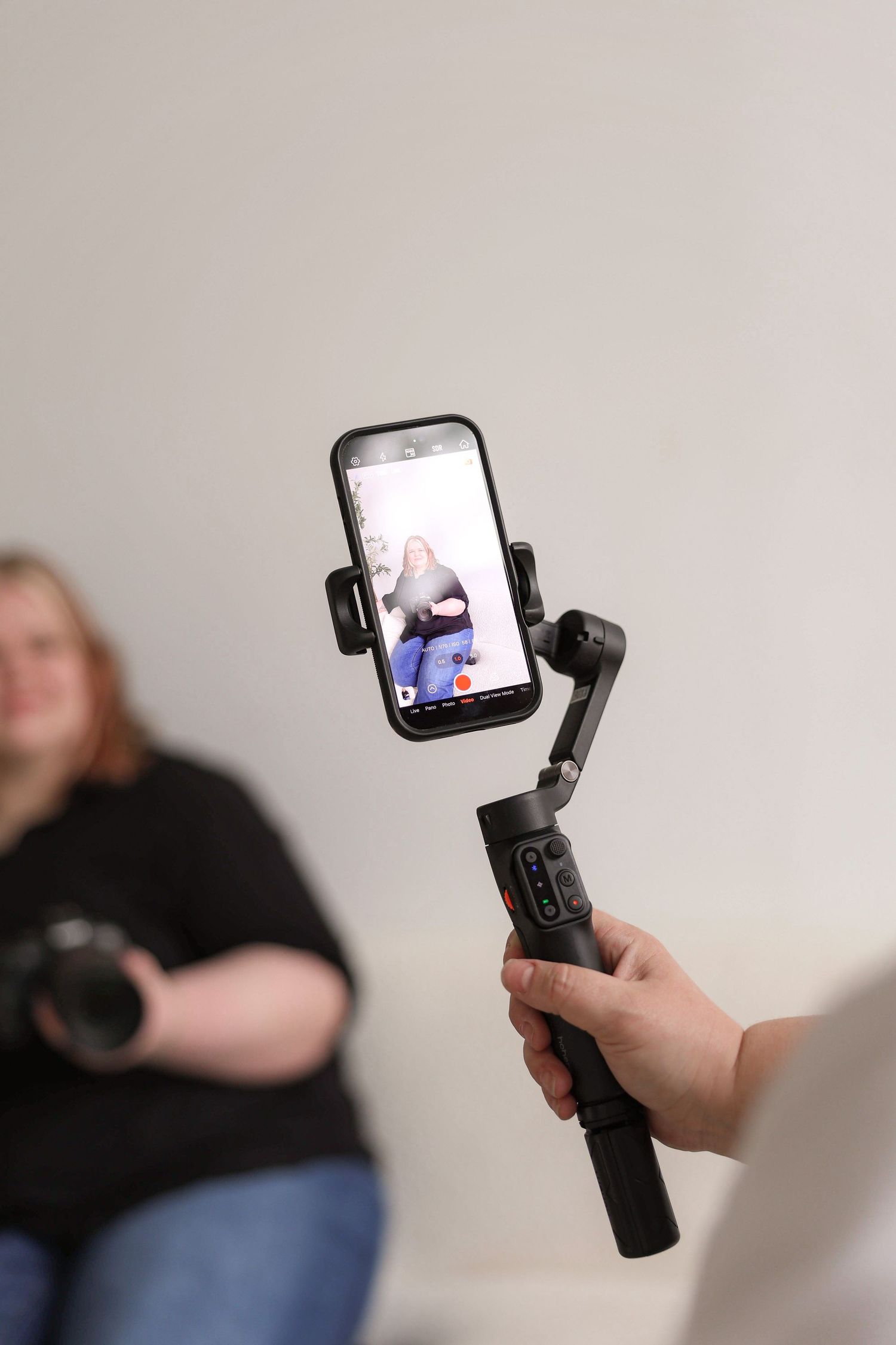 A handheld gimbal stabilizer holds a smartphone displaying content while someone films in a studio setting.