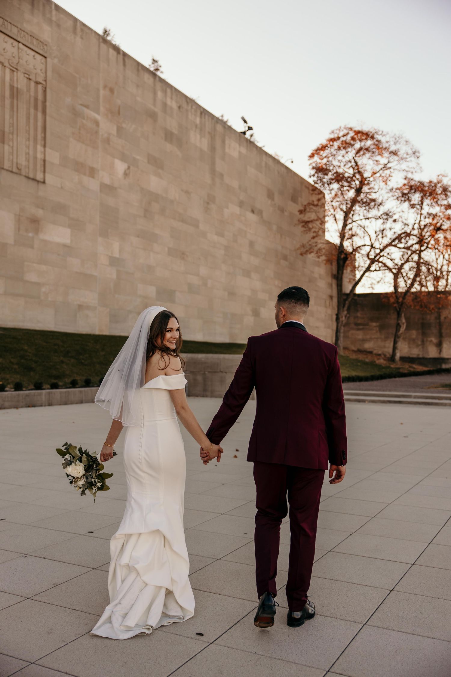 A bride and groom hold hands while walking along a modern stone building at sunset.