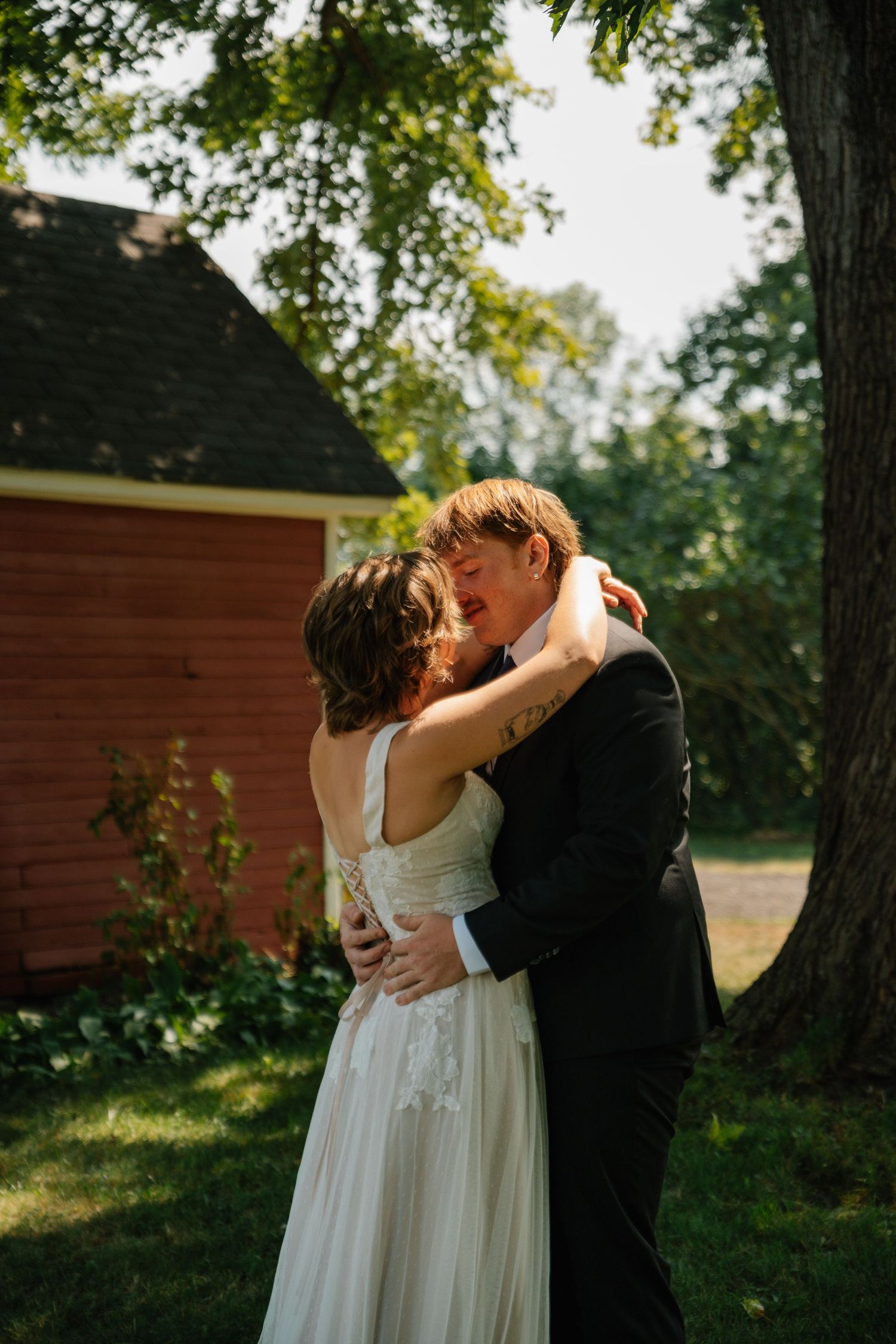 A couple in wedding attire share an intimate embrace in front of red barns during golden hour.