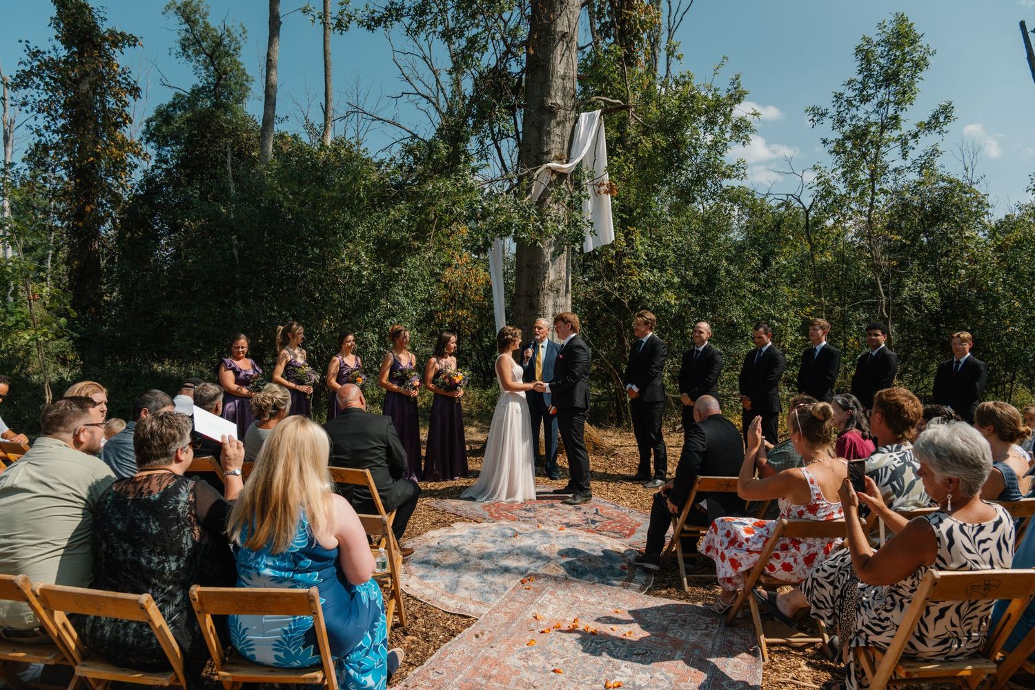 Outdoor wedding ceremony in a lush forest setting with bridal party in black attire and guests seated on wooden chairs.