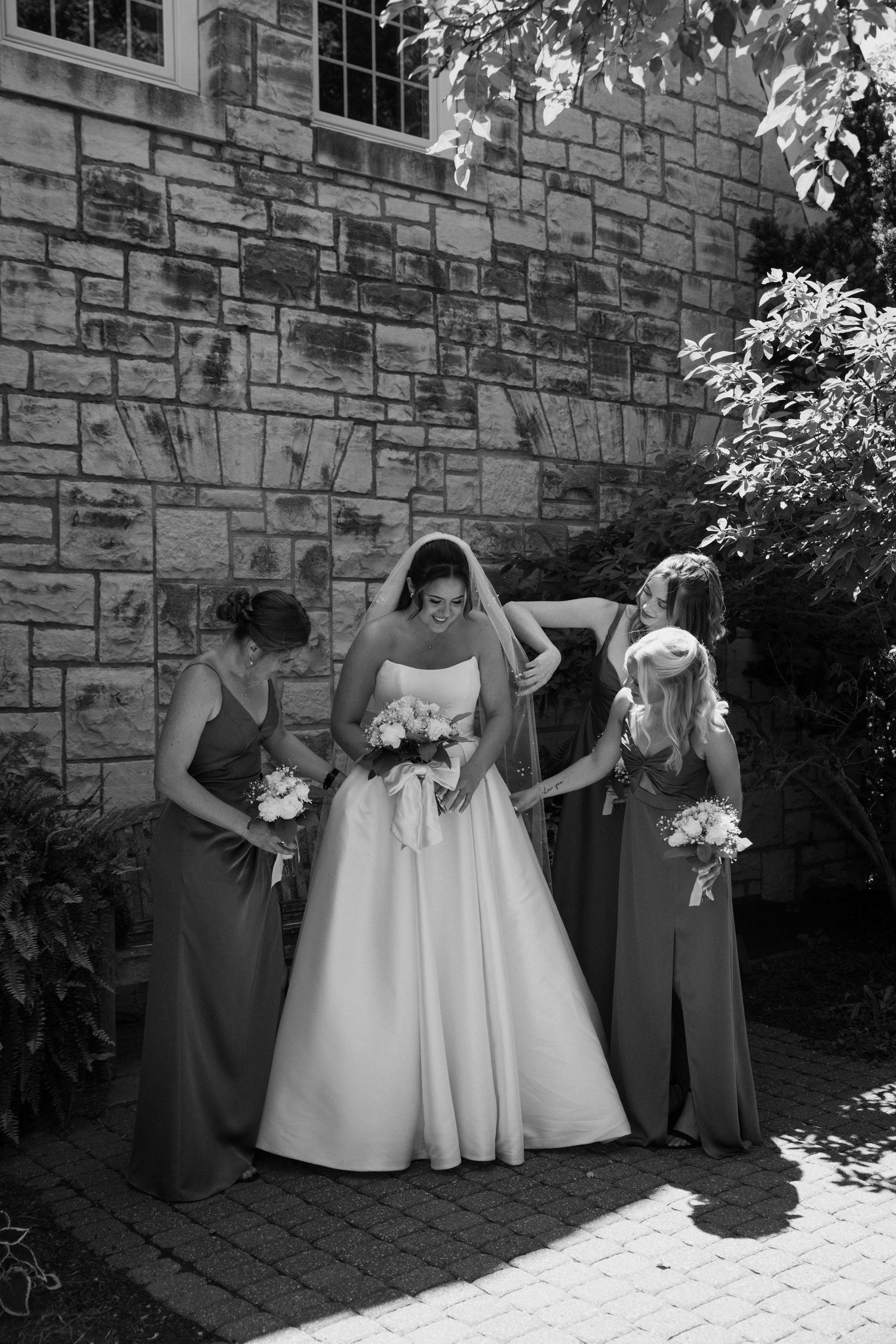 Bridesmaids in gray dresses pose with bouquets against a stone wall.