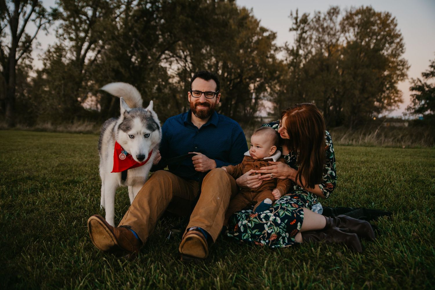A family and their husky dog relaxing together on grass during golden hour in a park setting.