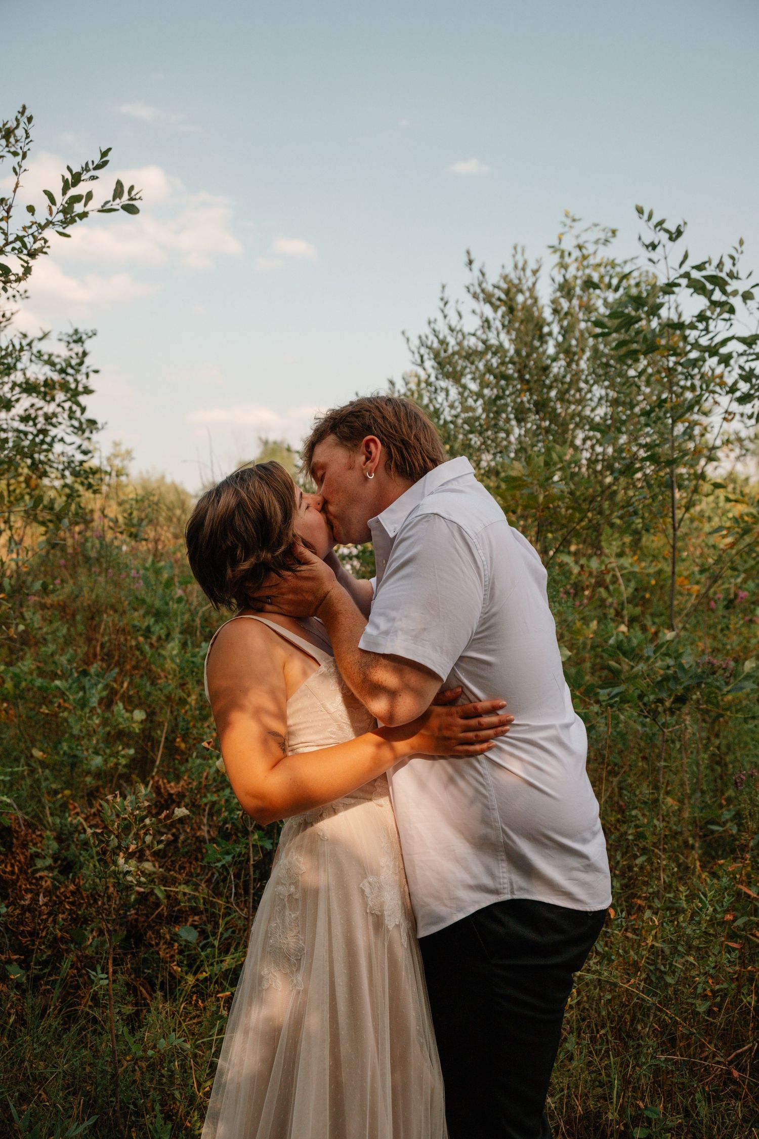 Couple sharing a romantic kiss outdoors in a lush green field, woman in white dress, man in light blue shirt.