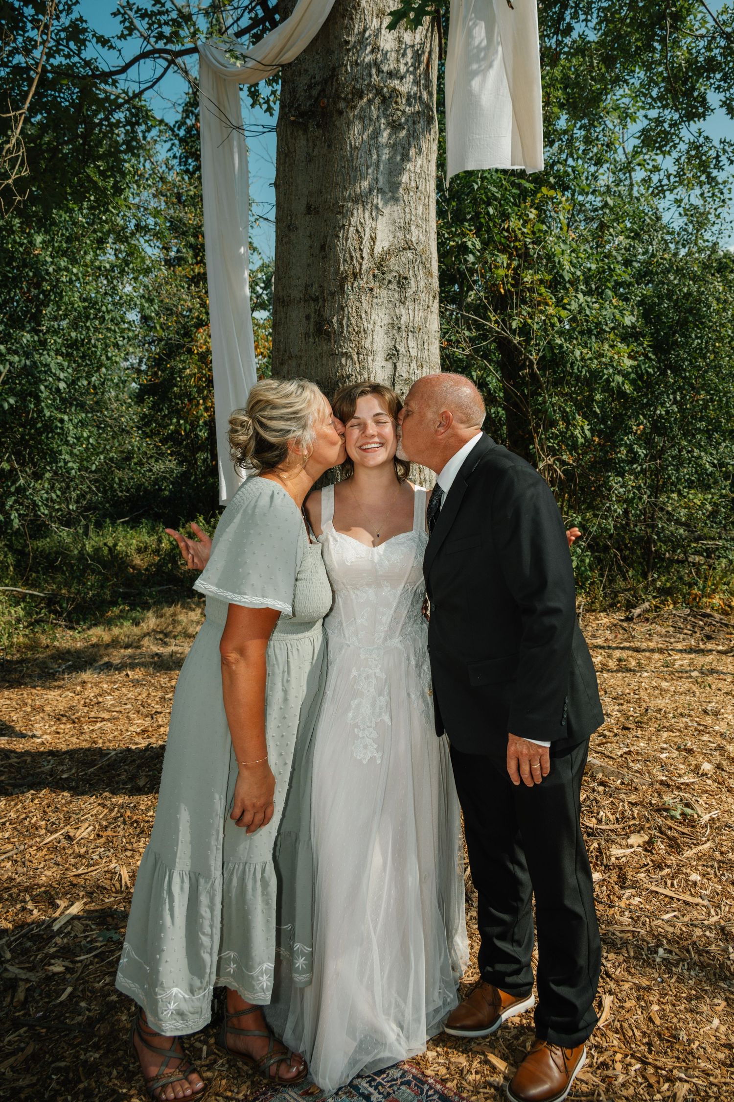 Bride in white gown receives kisses from parents outdoors at woodland wedding ceremony.