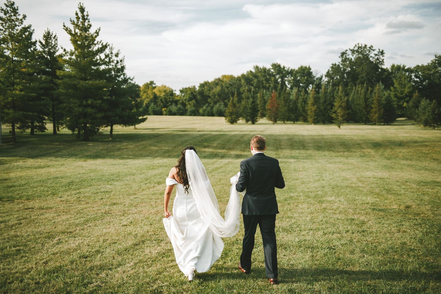 Newlyweds walk hand in hand across a scenic grassy field at sunset.