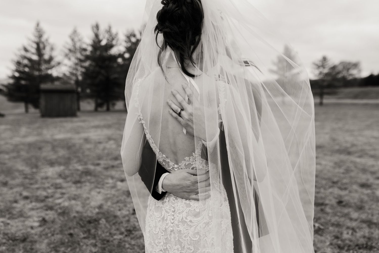 Back view of a bride's lace wedding dress and flowing veil in a rustic outdoor setting.