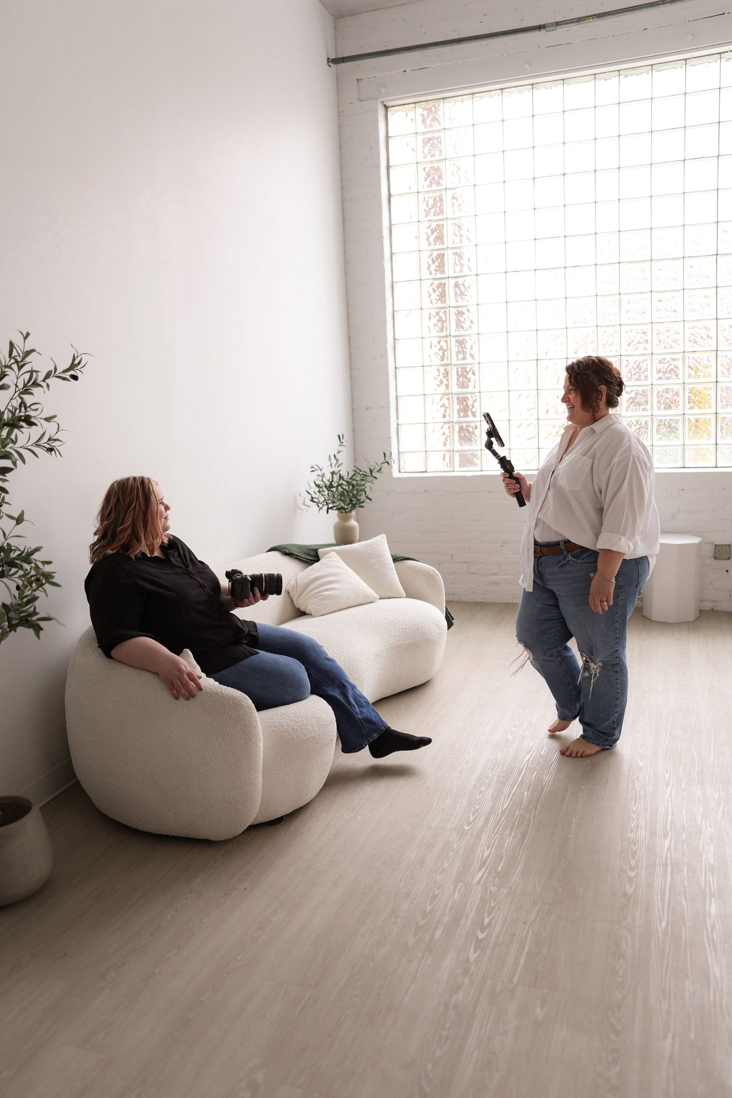 Two women in a bright white studio space, one seated on a modern sofa while the other holds a camera during a photo session.