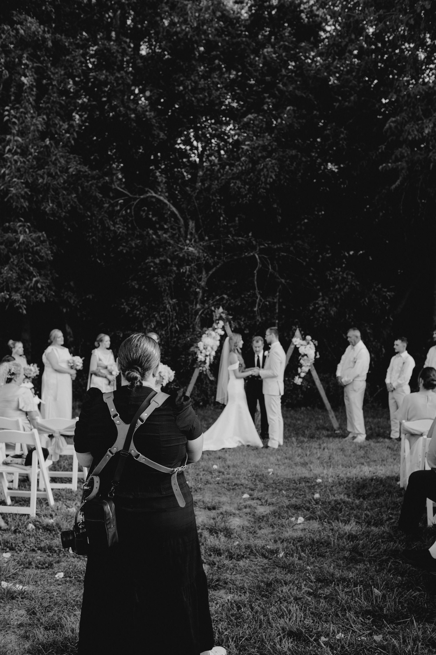 Black and white photo of an outdoor wedding ceremony with guests seated in white chairs.