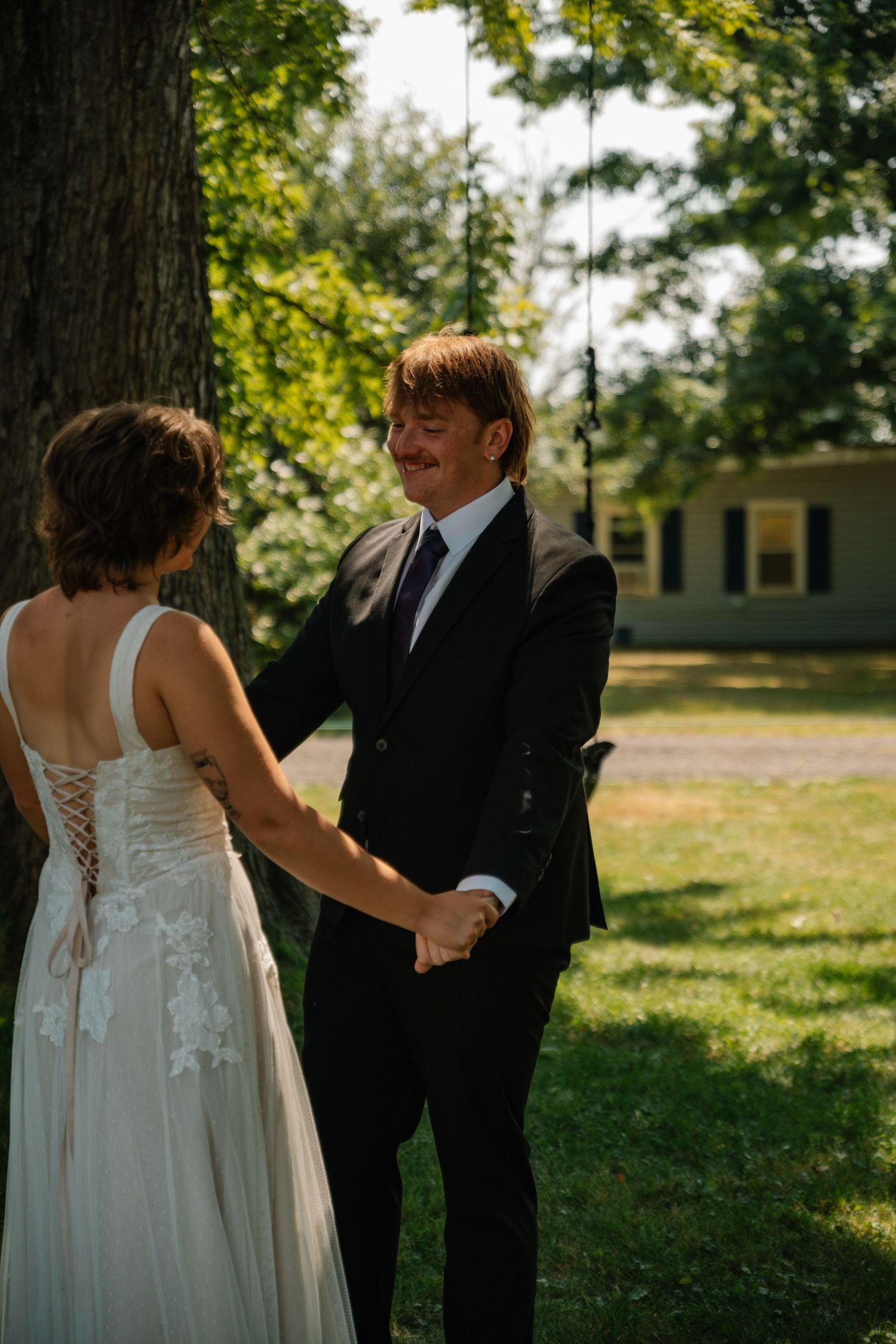 Groom in black suit smiling joyfully while holding hands with bride in lace wedding dress outdoors under a large tree.