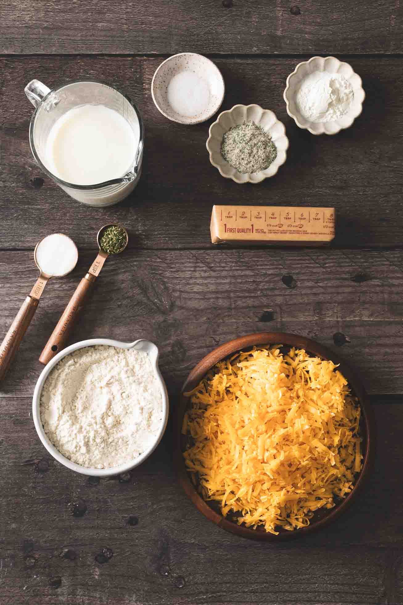 Baking ingredients including milk, flour, shredded cheese and measuring spoons arranged on dark wooden surface.
