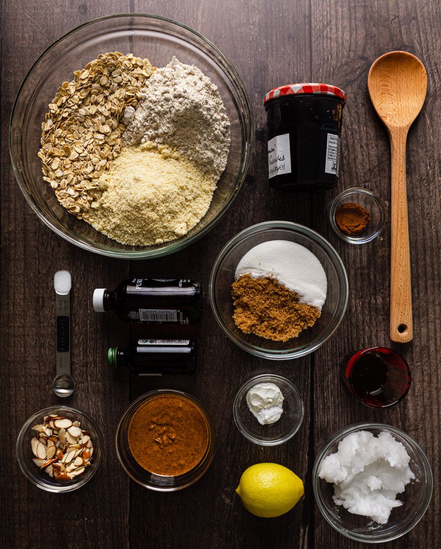 Baking ingredients arranged on dark wood table include flour, spices, coconut oil, lemon, jam and wooden spoon.
