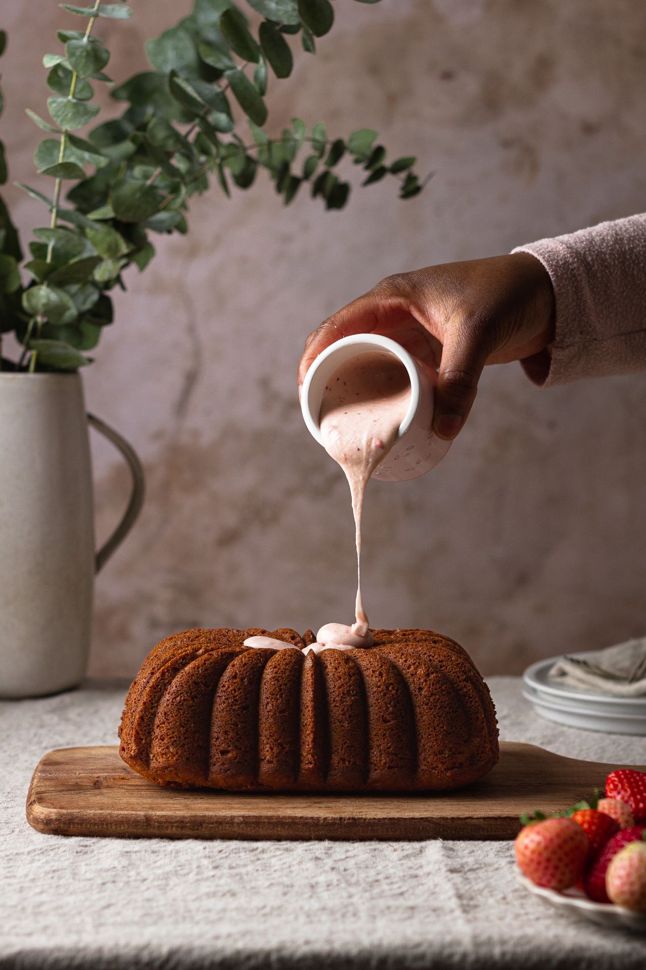 Pouring glaze over freshly baked bundt cake with eucalyptus and strawberries in background.