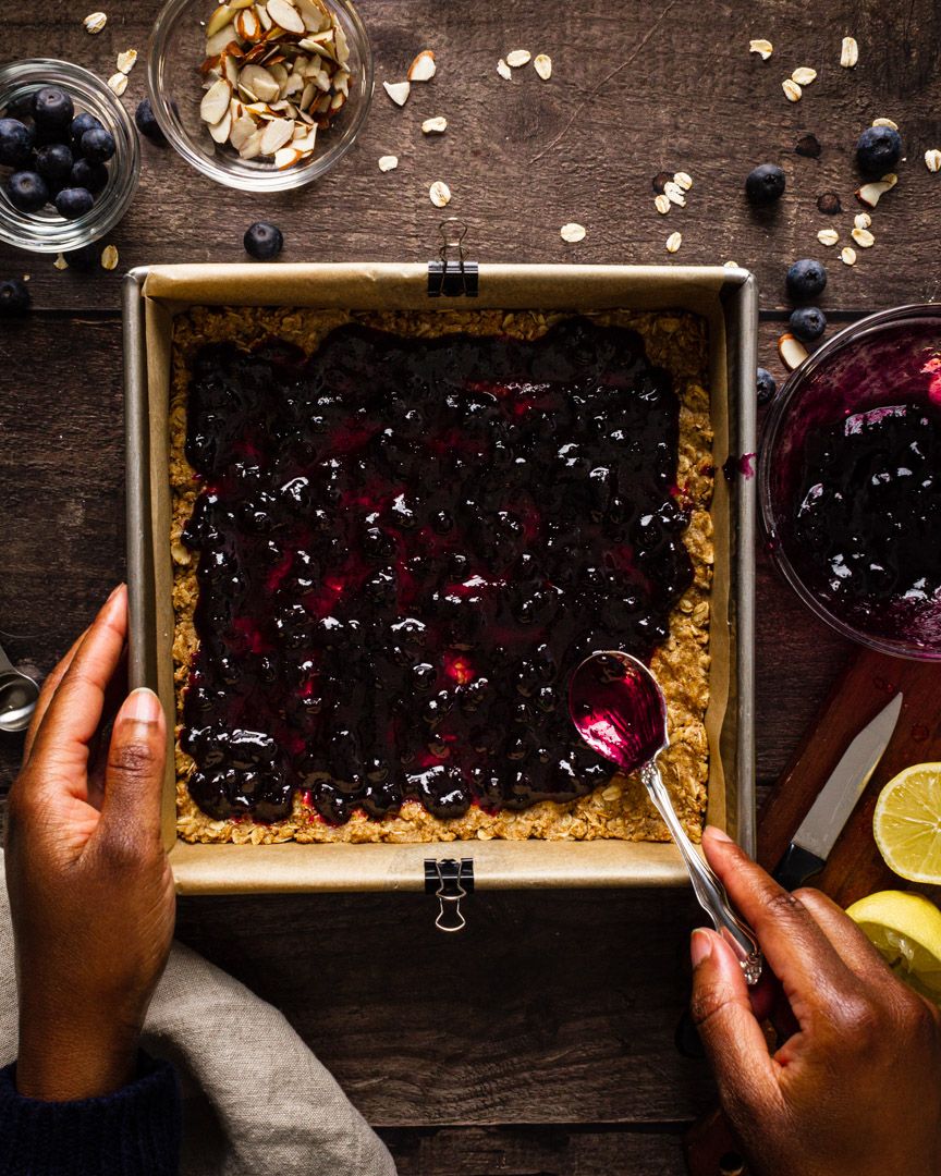 Overhead view of hands preparing a square blueberry tart on wooden table with nuts and citrus garnishes.