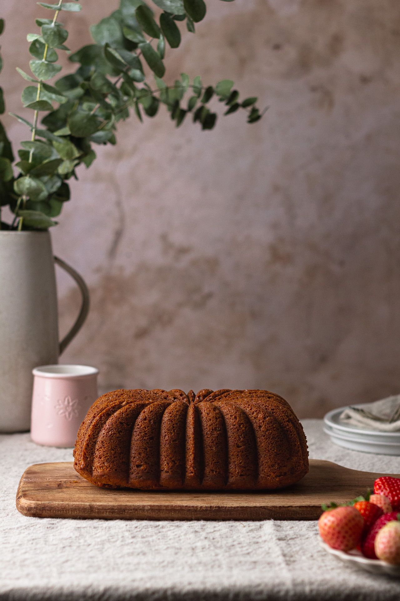 Homemade bundt cake on wooden board with eucalyptus and fresh strawberries in rustic setting.