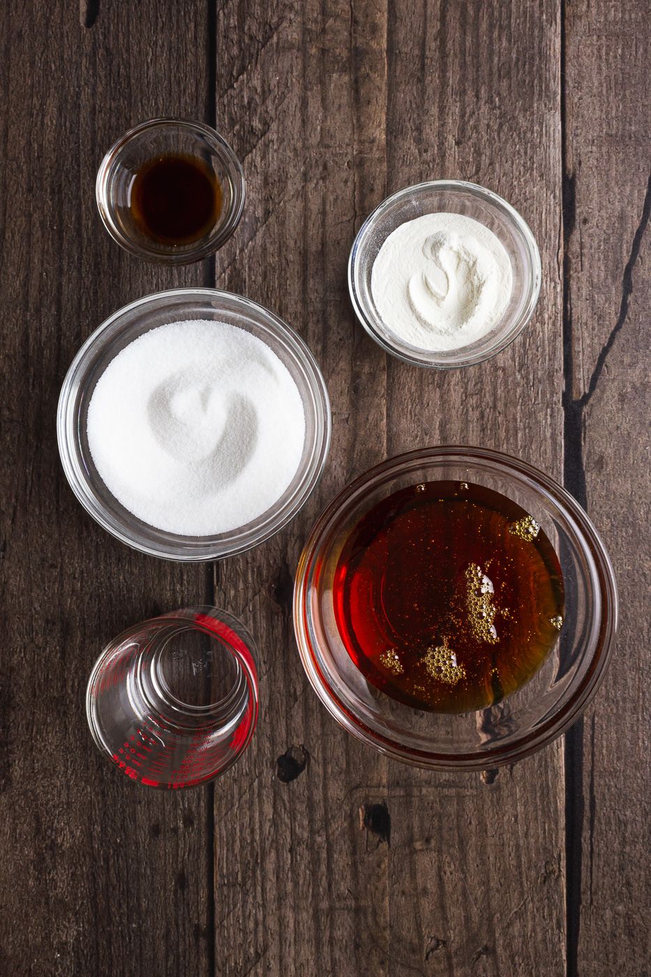 Overhead view of glass bowls with sugar, honey, and other baking ingredients on a rustic wooden table.