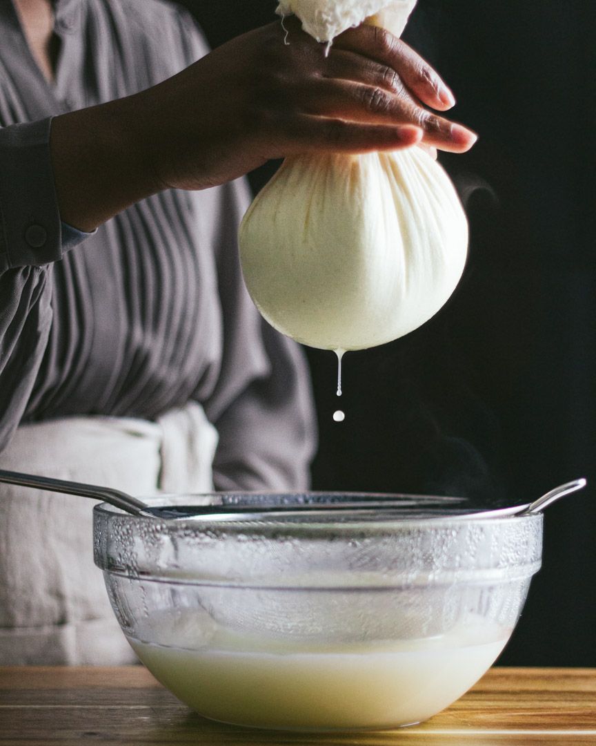 Hands strain homemade ricotta ch through cheesecloth over a glass bowl, with white liquid dripping into the collection bowl.
